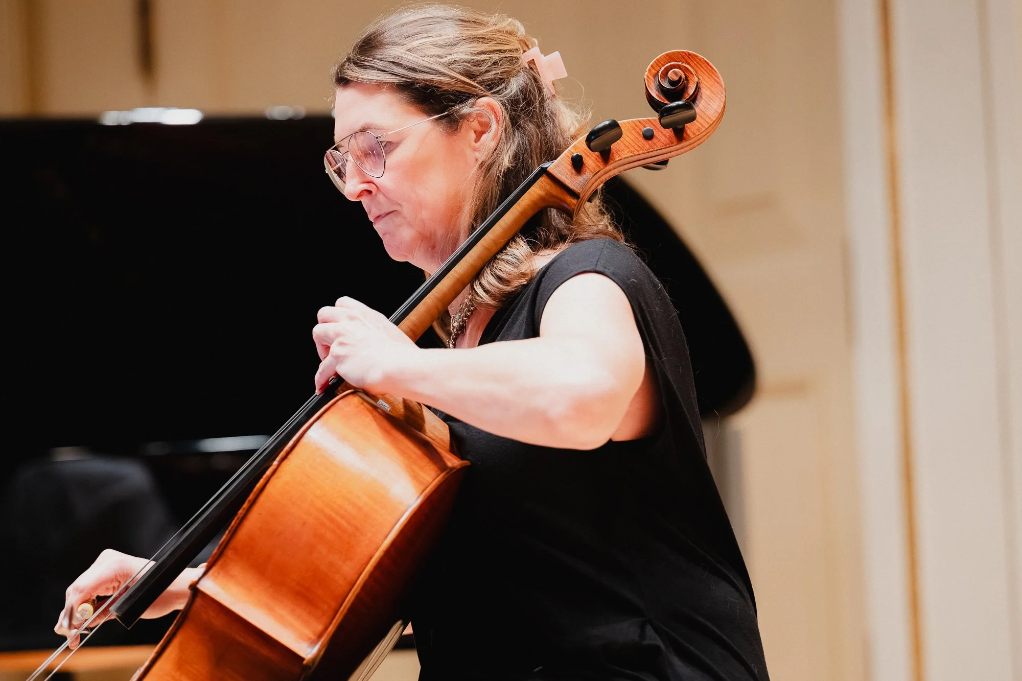 Female cellist in profile wearing glasses and a black top, playing with full bow extension, the scroll of the cello prominent above her shoulder, in Weill Recital Hall