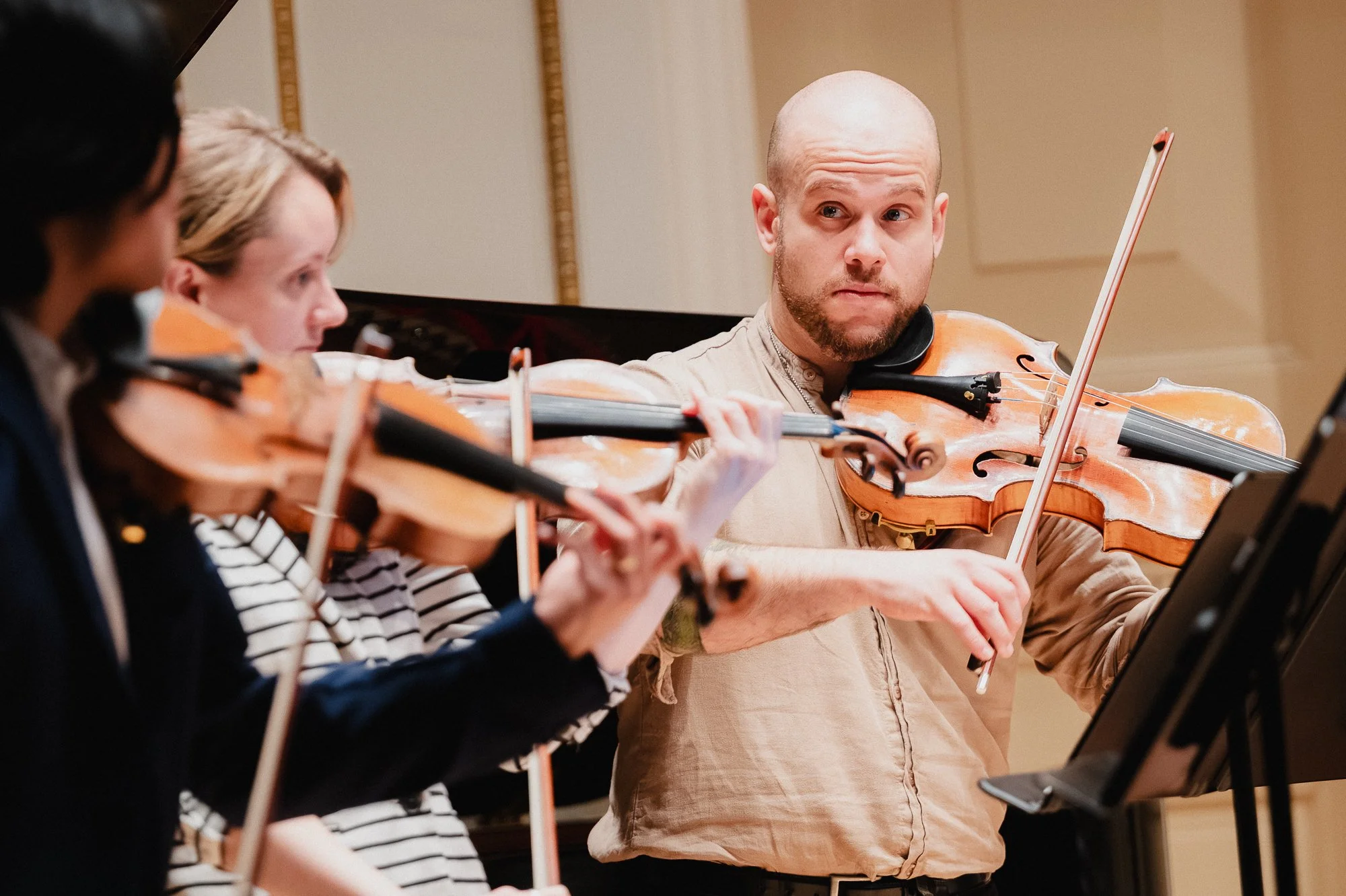 Three string players with violins and viola mid-rehearsal in Weill Recital Hall, the bearded bald violist in the background looking directly across at the other players while playing