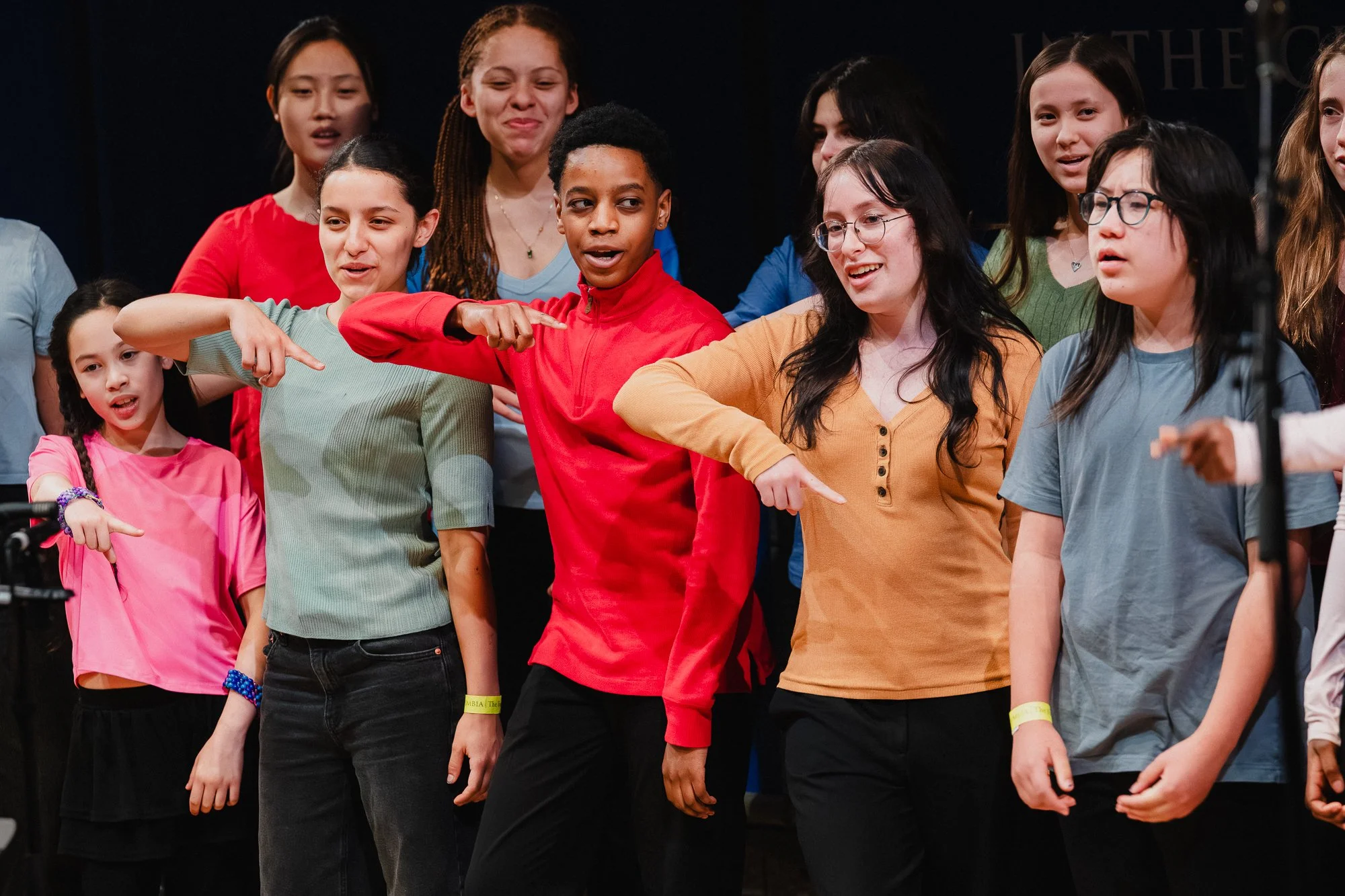 Group of youth singers on stage mid-performance, several pointing outward in unison, wearing colorful tops including red, green, and yellow against a dark background with other performers behind them