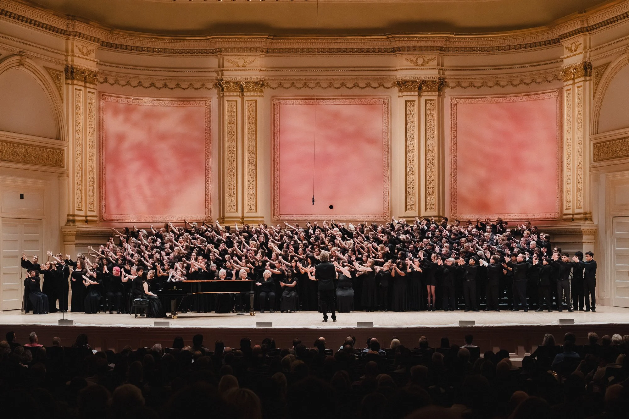 The full combined choir in black fills the Carnegie Hall Stern Auditorium stage, with Eric Whitacre nducting from center-front, arms extended, grand piano stage left, three large salmon-toned panels behind the ensemble.]