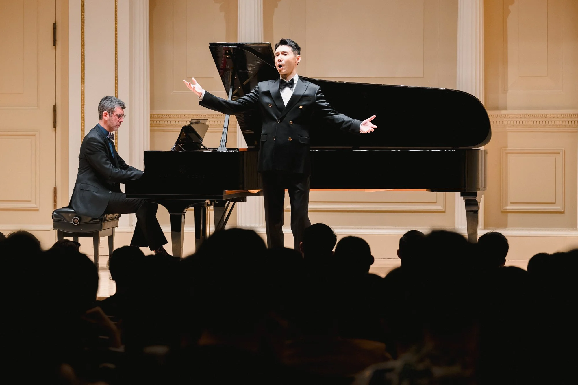 Tenor in black tuxedo performing with arms extended alongside pianist accompanist at Weill Recital Hall, audience silhouettes in foreground.