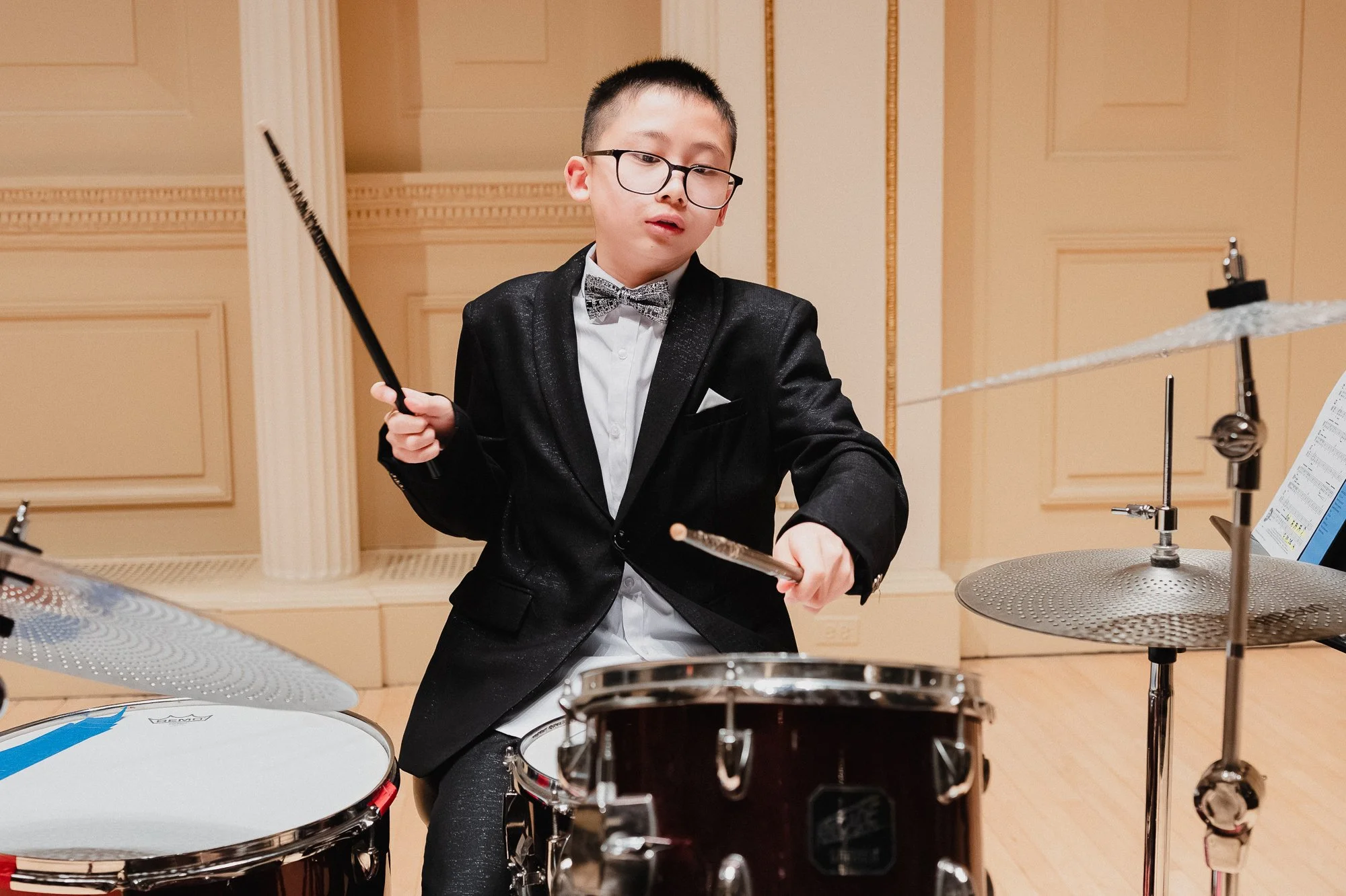 A young boy in a black tuxedo jacket and bow tie plays a full drum kit on stage, both sticks moving, expression focused and intent