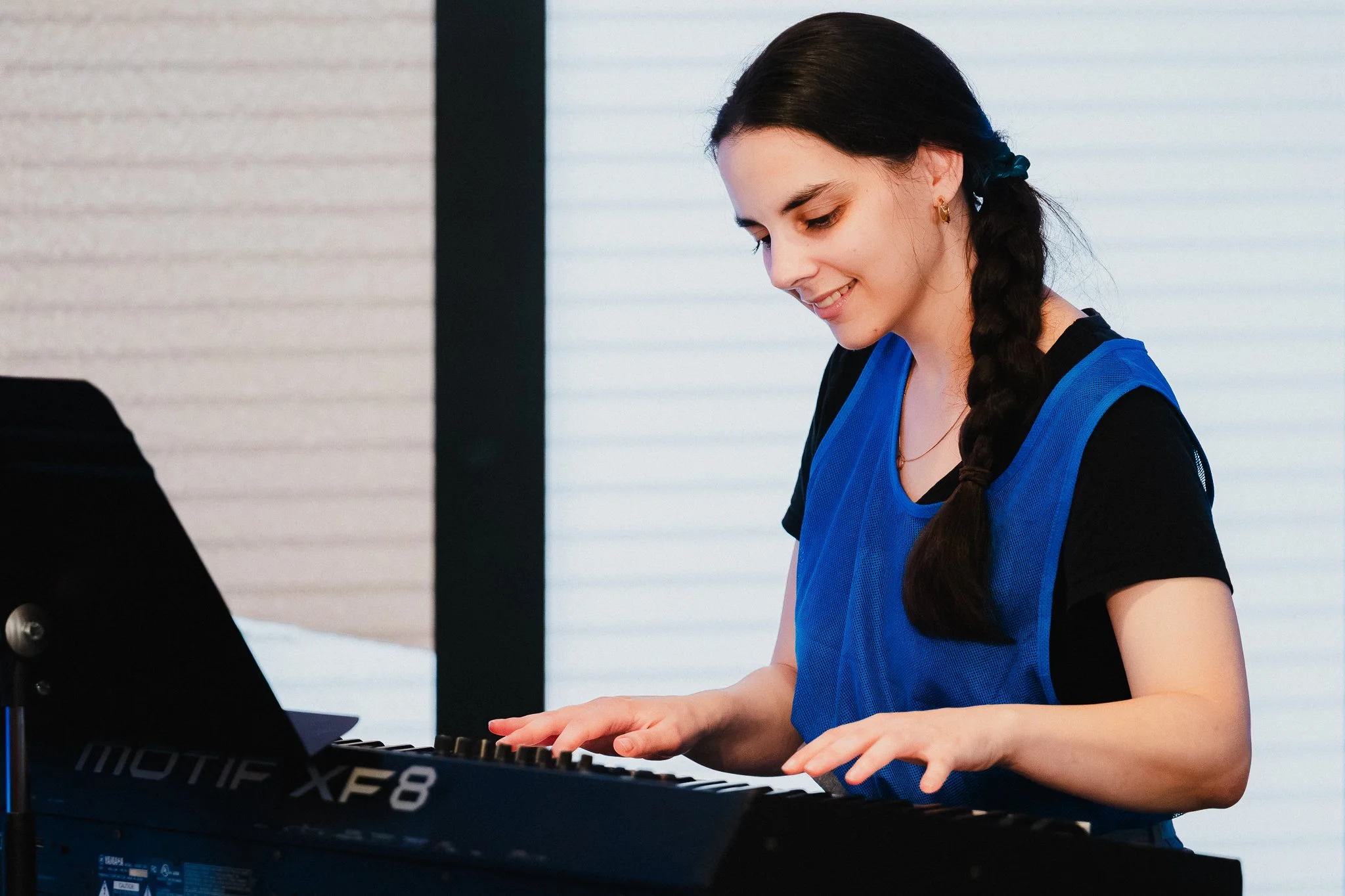 Composer Maria Caputo plays a keyboard, head slightly bowed, smiling, wearing a blue mesh vest over a black shirt