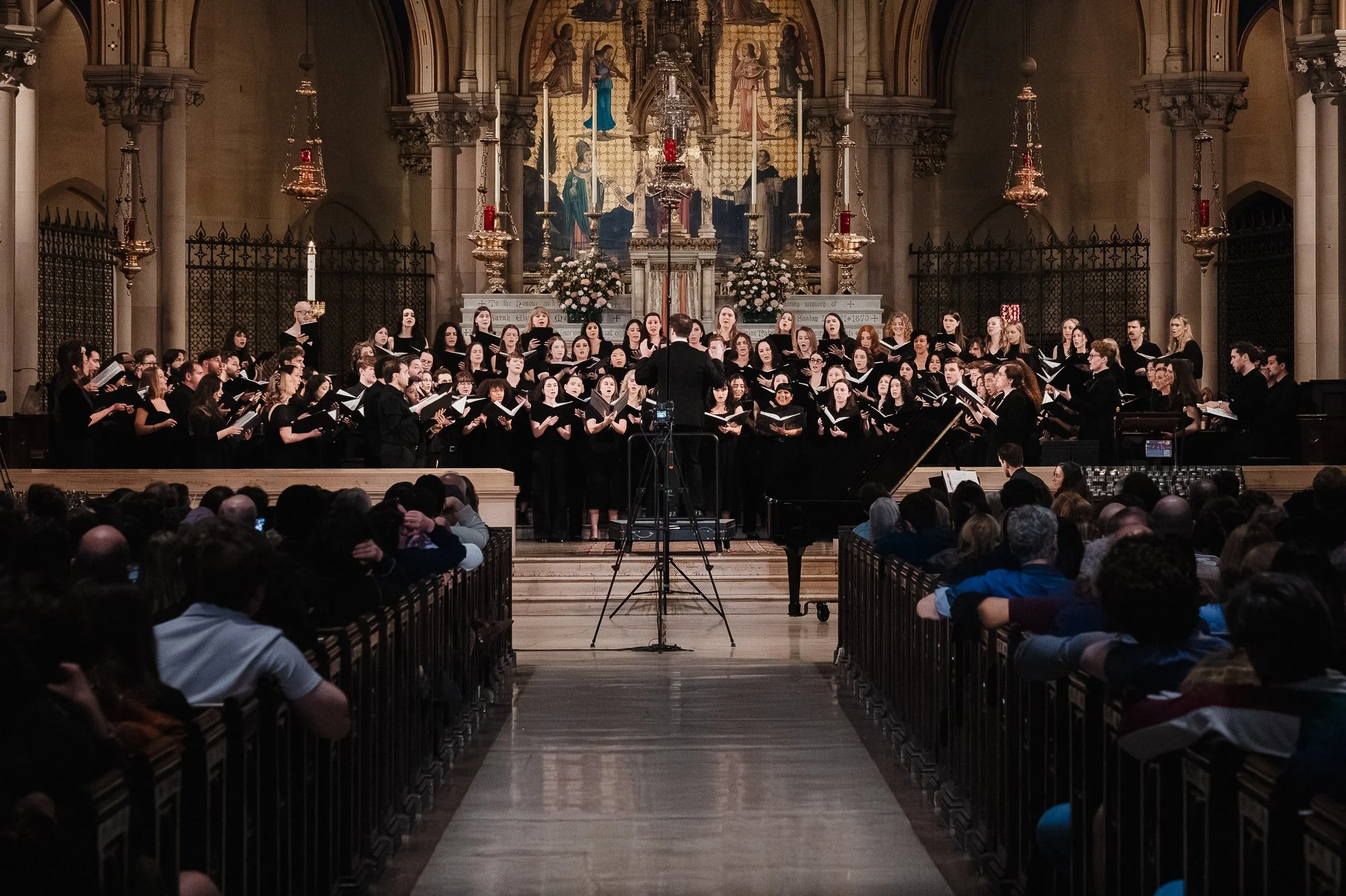 Full choir in black filling the chancel of St. Mary the Virgin, conductor facing away from camera at a podium with camera rig, grand piano to the right, ornate altar and reredos lit behind the ensemble, audience in foreground pews