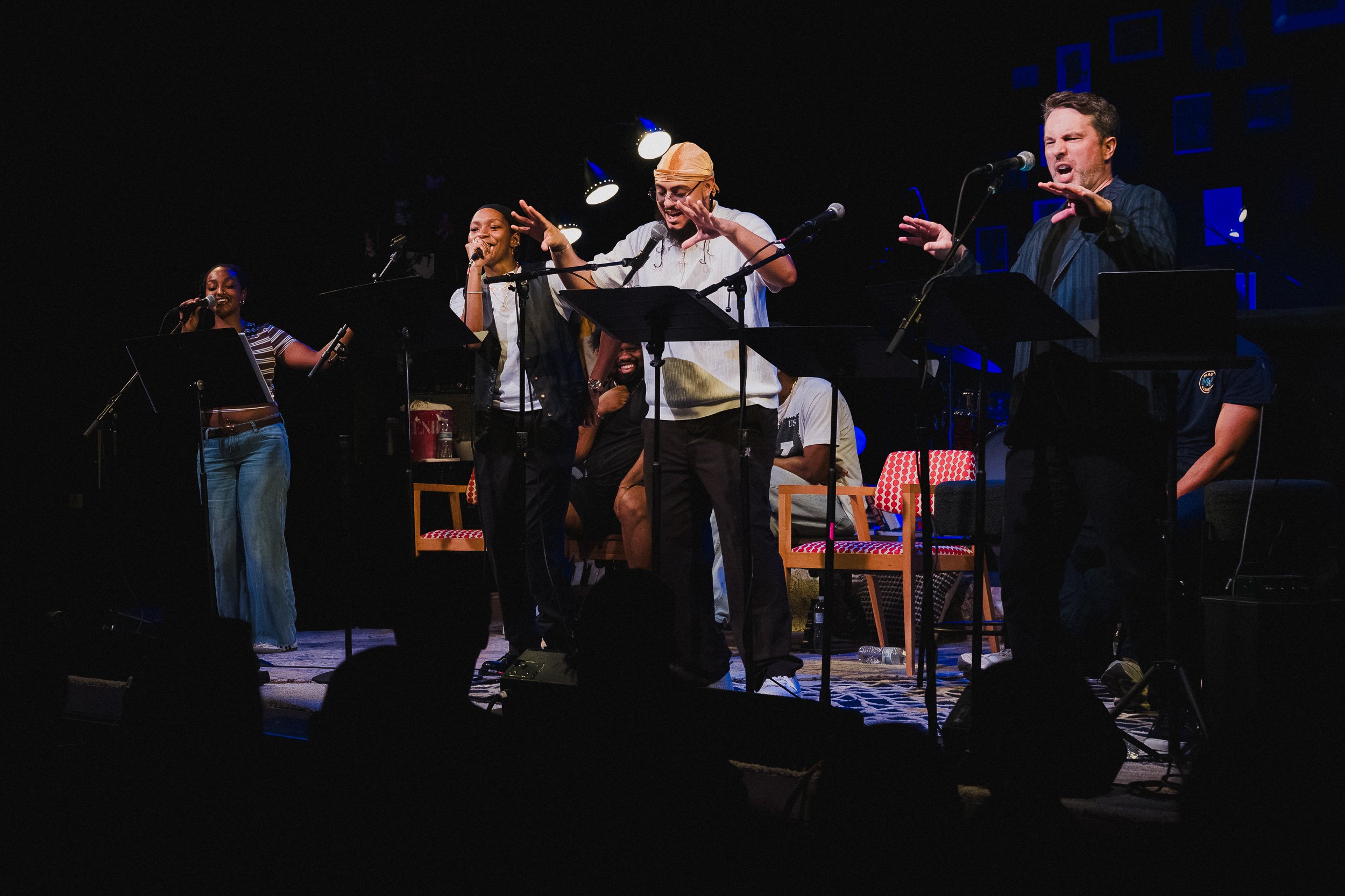 Wide view of five BLUDLINE performers at mic stands under blue stage light at Greenwich House Theater, arms raised
