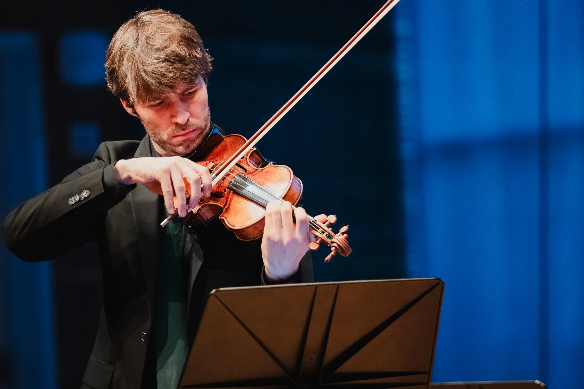 A violinist in a black suit plays with his bow arm extended on a downstroke, eyes lowered toward a music stand, blue-lit windows behind him.