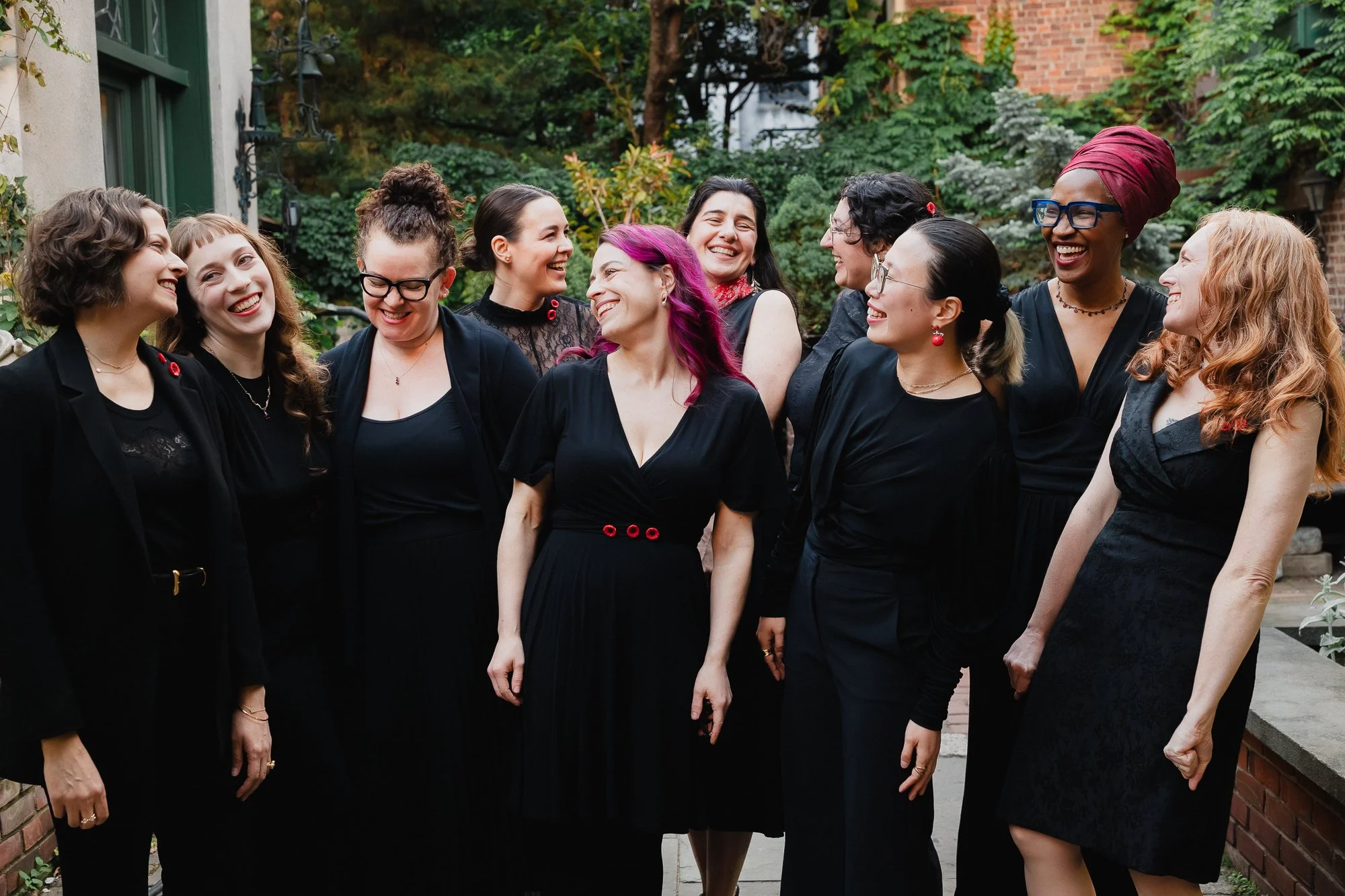 The full ensemble in black attire standing together in a garden courtyard, smiling, surrounded by green foliage and brick walls