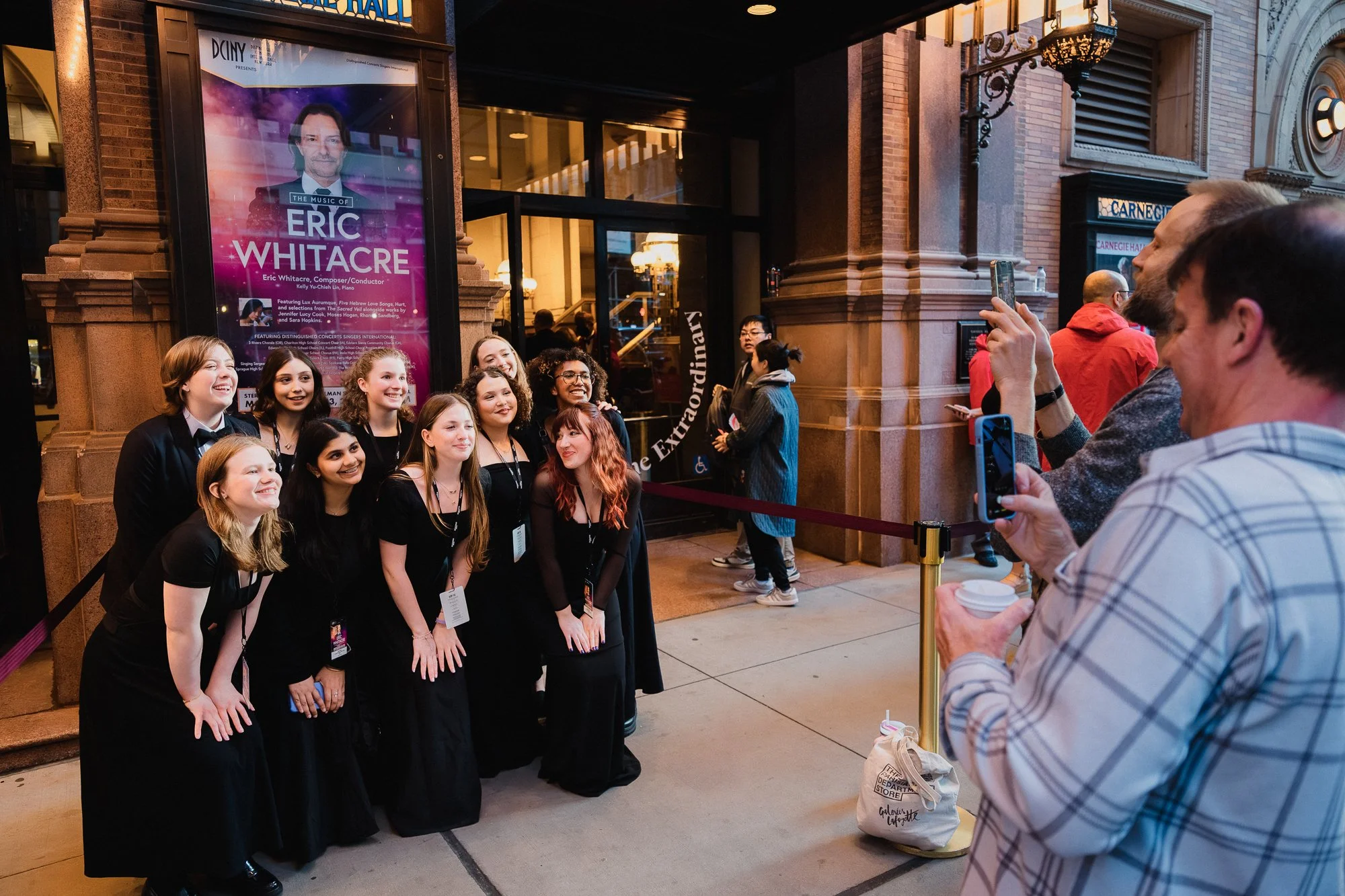 Young women in black concert dress pose in front of an Eric Whitacre event banner on the Carnegie Hall exterior while a man photographs them with a phone, evening light, sidewalk crowd behind.
