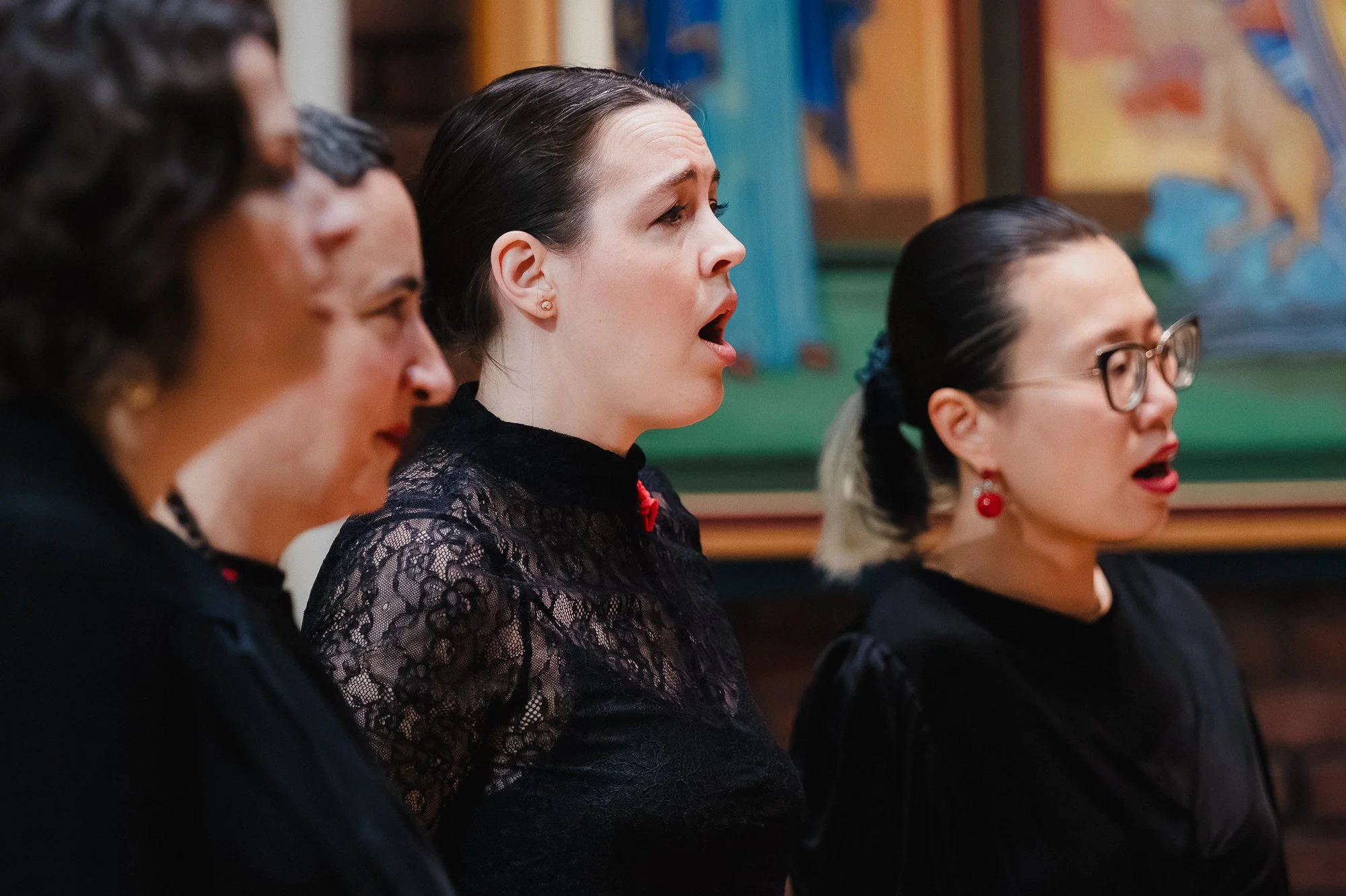 Three women singing in profile in a brick church, the central singer in a black lace top with a red pin, a colorful triptych altarpiece visible behind them