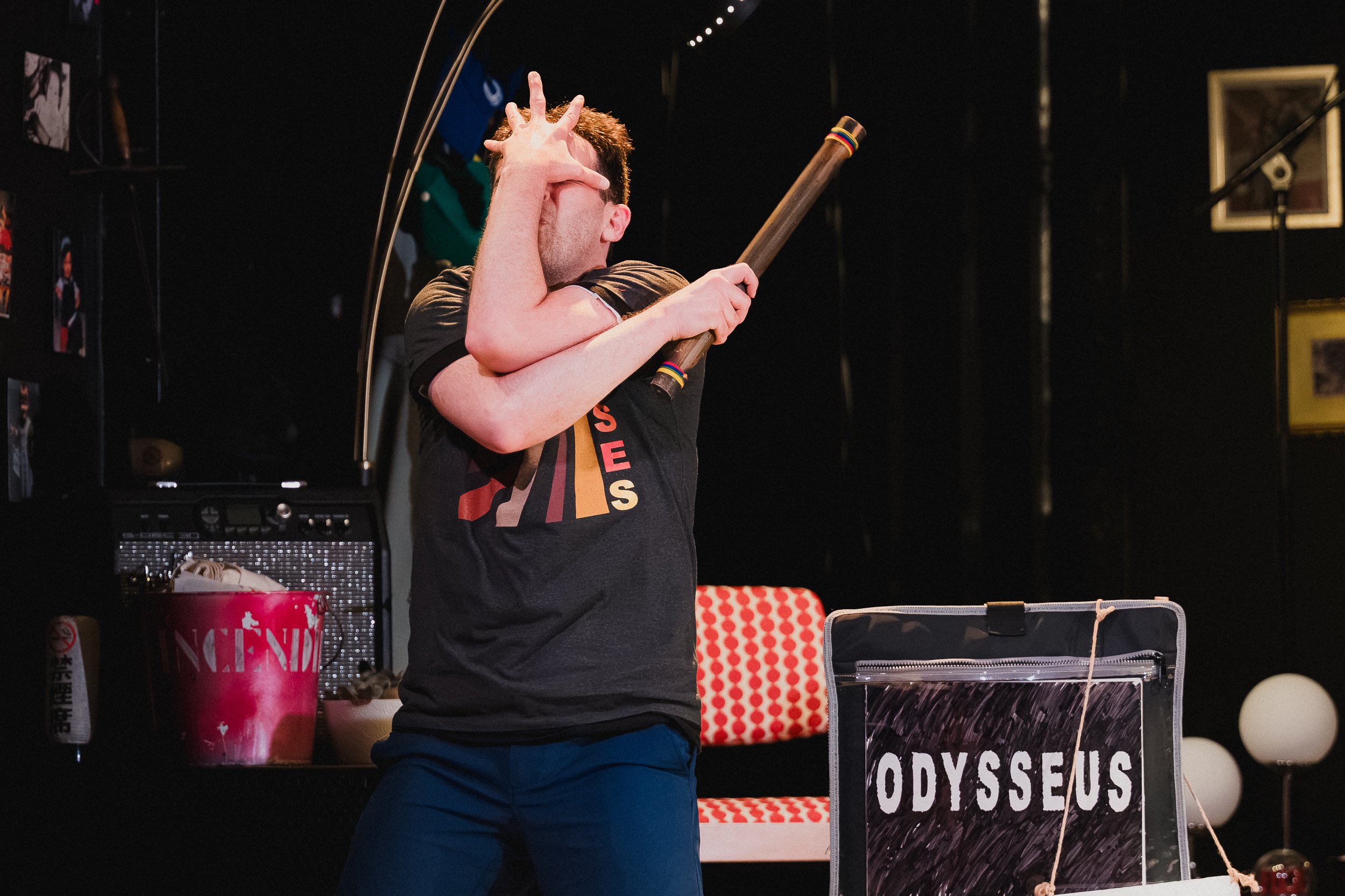Joseph Medeiros grips a wooden staff across his body, face turned away, on the small stage at Greenwich House Theater