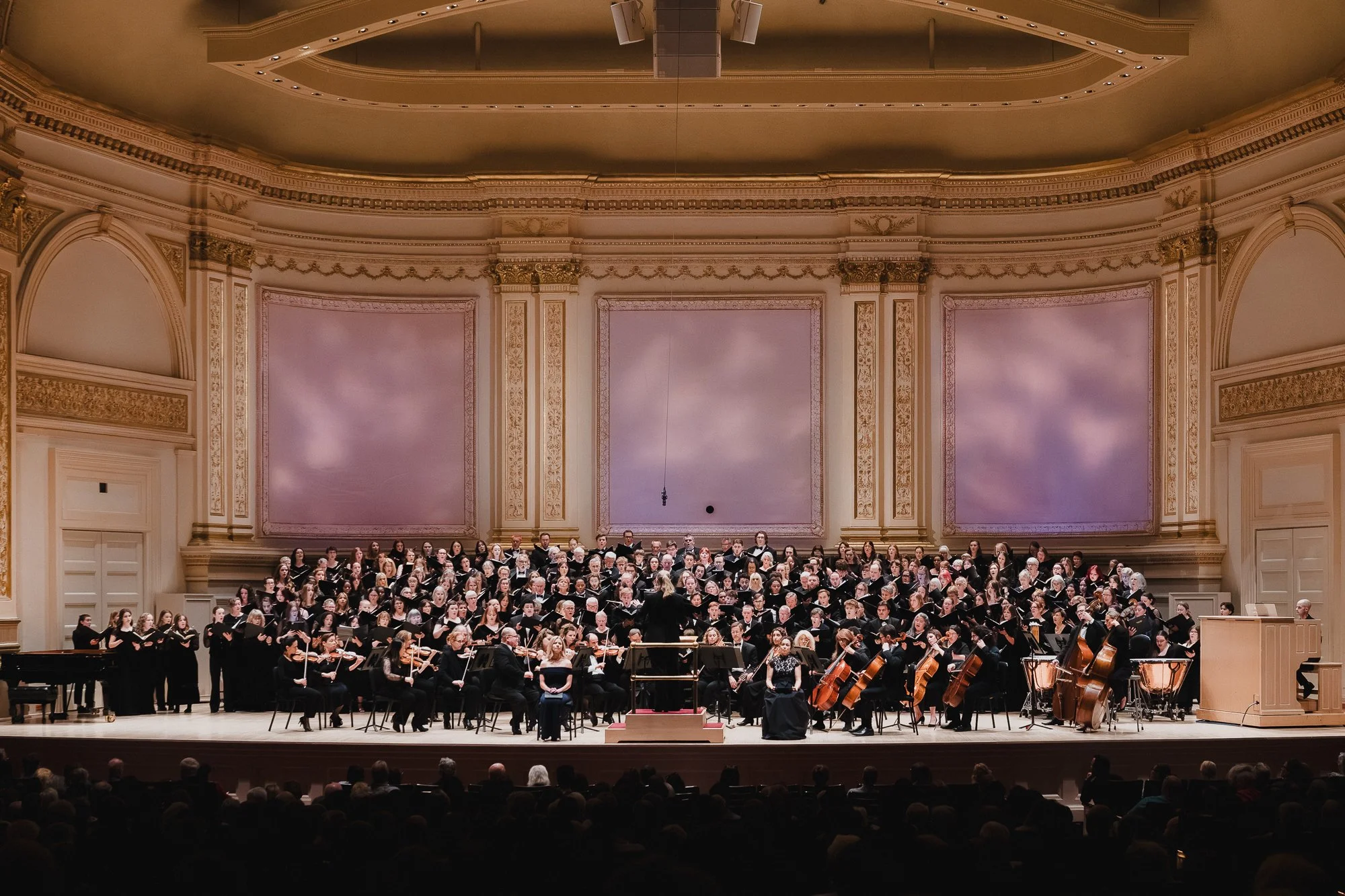 Wide view of Stern Auditorium from the rear of the house, full choir and orchestra on stage against three large pink-lit panels, ornate gold ceiling and arched side walls visible