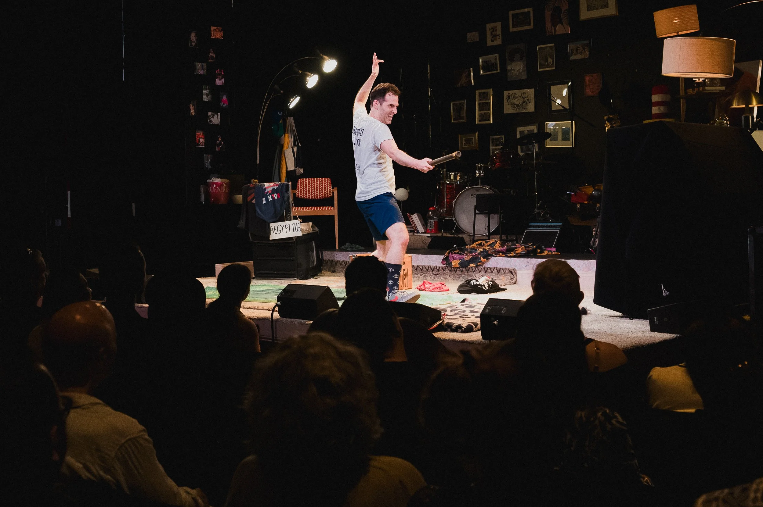 Joseph Medeiros stands on a raised platform with a microphone, one arm up, audience silhouetted in the foreground at Greenwich House Theater