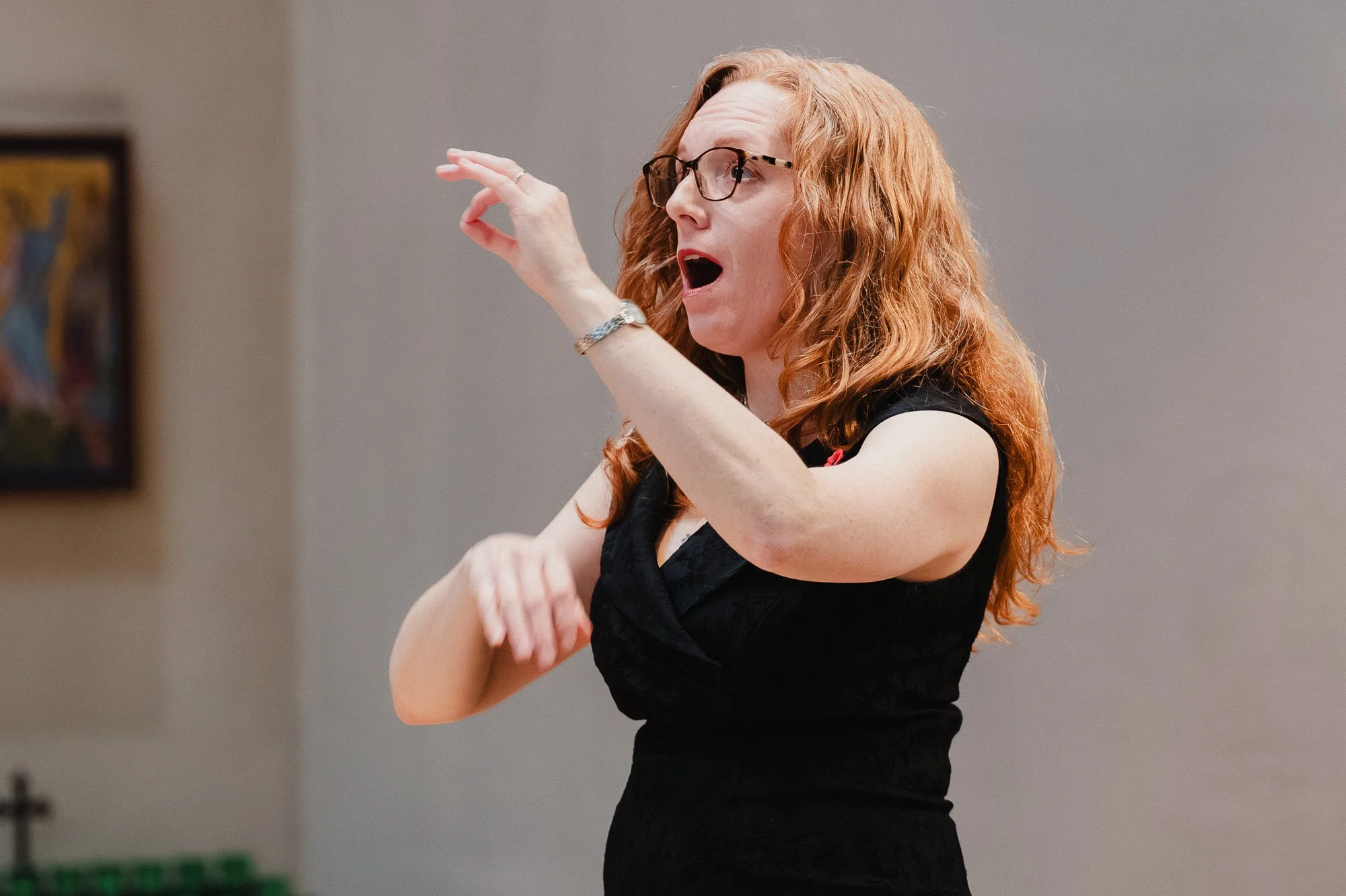 A conductor with long red hair and glasses in a black dress, both hands raised mid-gesture, mouth open as she sings with the ensemble, an icon on the wall behind her