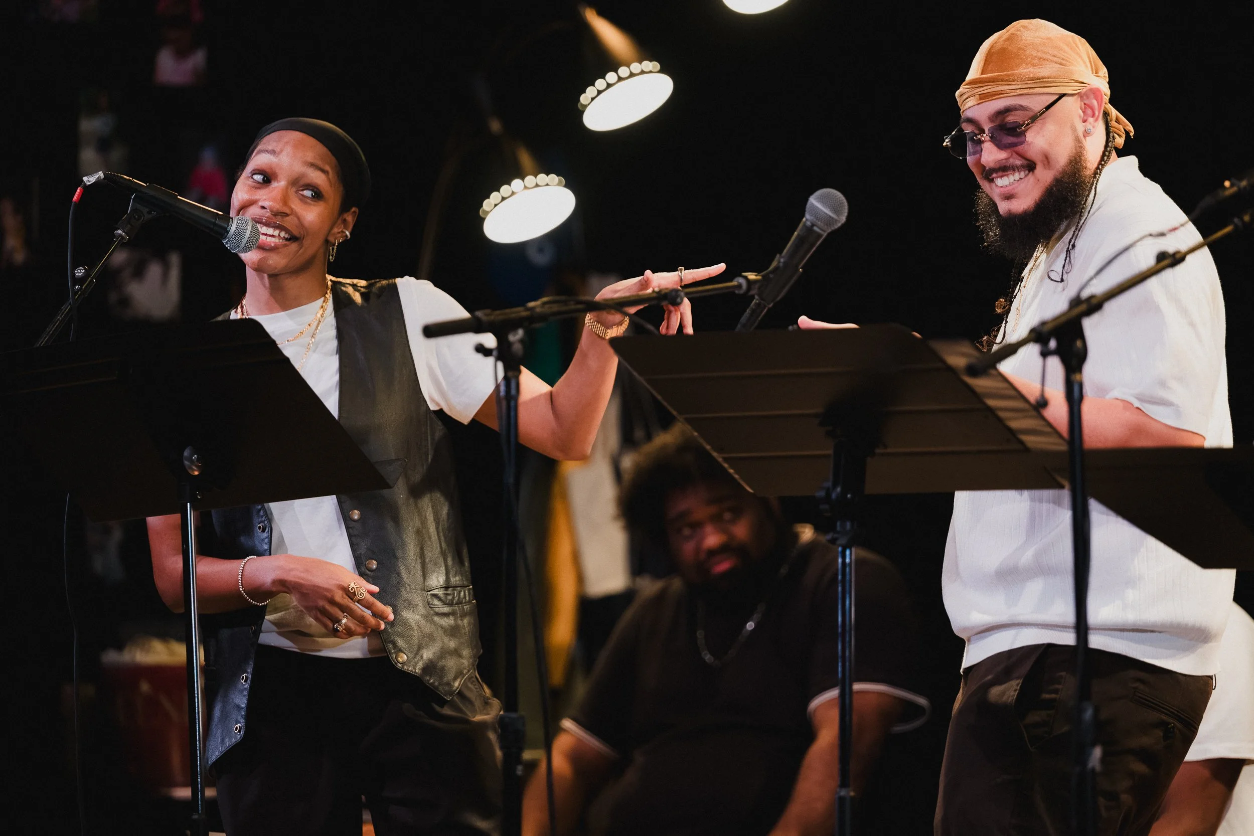 Two BLUDLINE performers at mic stands mid-exchange, one gesturing toward the other, a third seated behind, at Greenwich House Theater