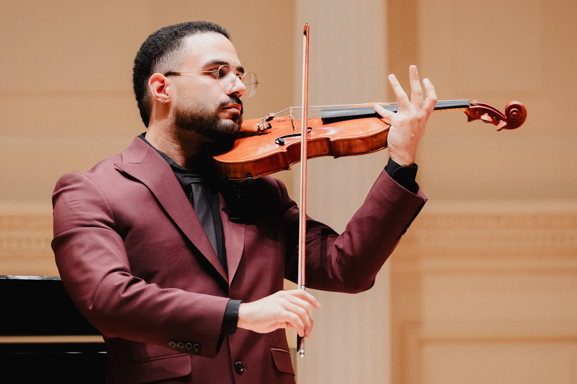 A man in a dark burgundy suit plays violin in close-up, bow raised vertically, eyes closed, expression absorbed, Carnegie Hall stage in soft focus behind