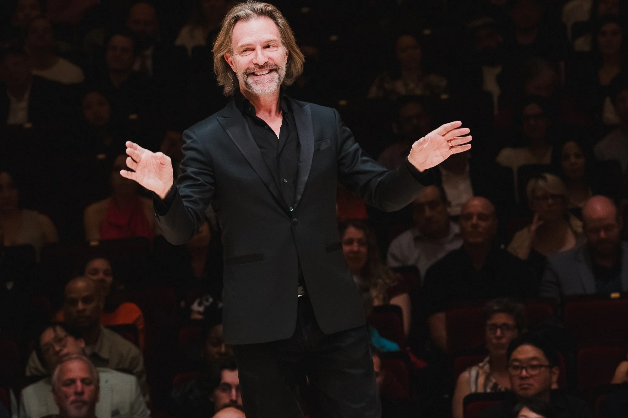 Eric Whitacre smiling broadly with both arms open wide, facing the full audience at Carnegie Hall, the warm-lit house visible behind him.