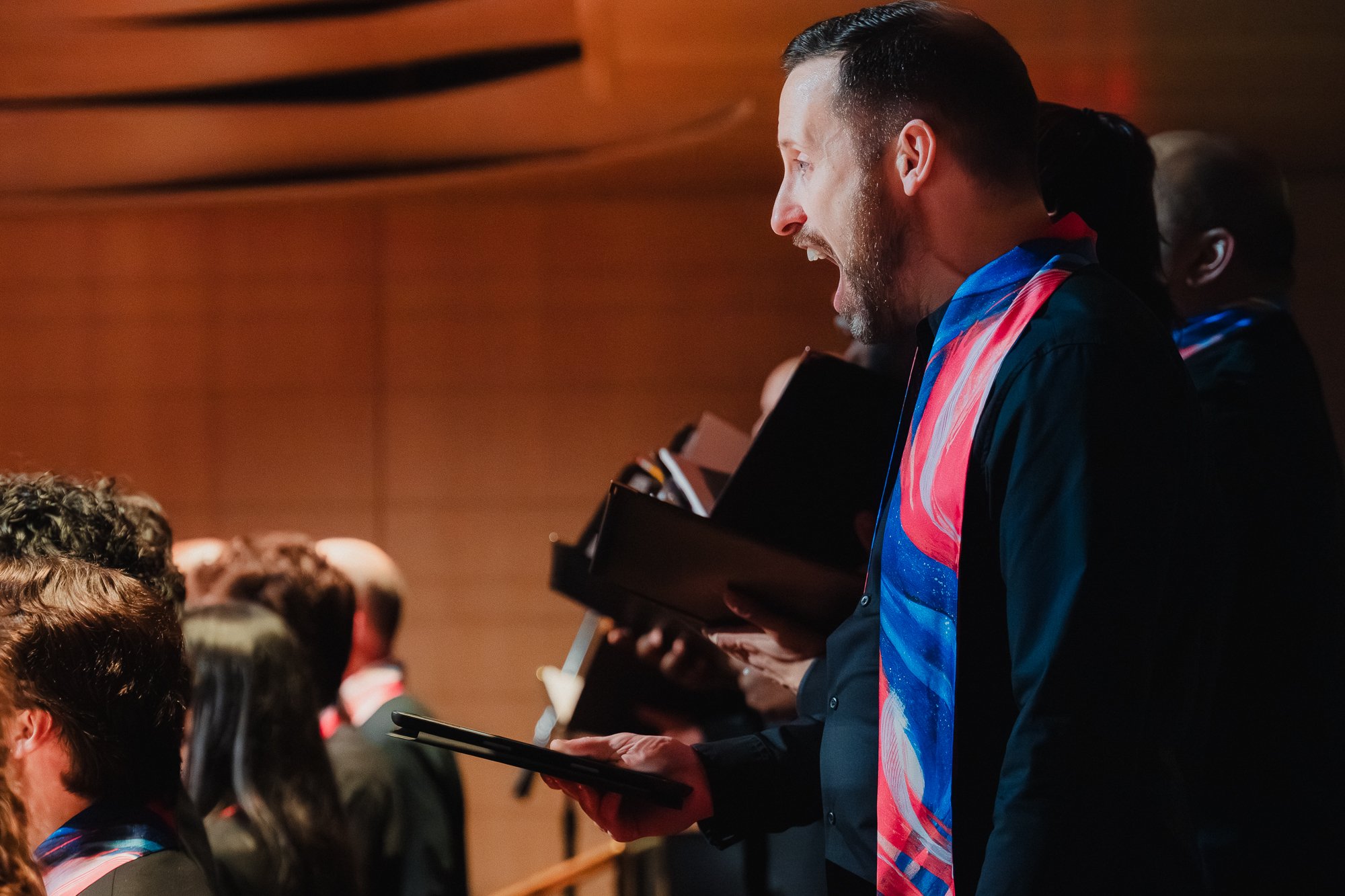 Close profile of a male singer with dark hair and a beard, mouth wide open mid-phrase, wearing black with a bold blue, pink, and red scarf, holding a tablet, other choir members slightly out of focus behind him