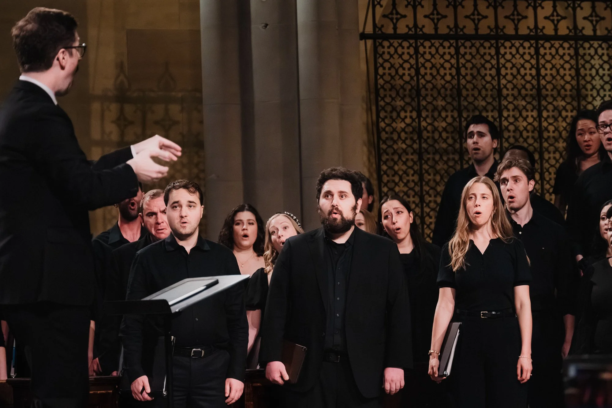 Conductor seen from behind at left, right hand extended mid-gesture toward choir, group of mixed singers in black at center and right, bearded male singer prominent at center foreground, ornate gilded choir screen behind