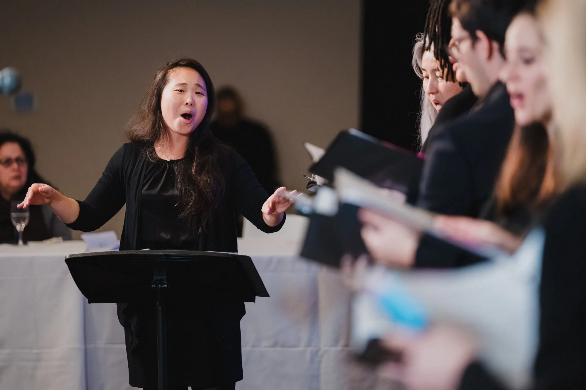 Conductor in black leads the Teachers College Choir at United Nations headquarters, singers with folders visible in soft focus at right.