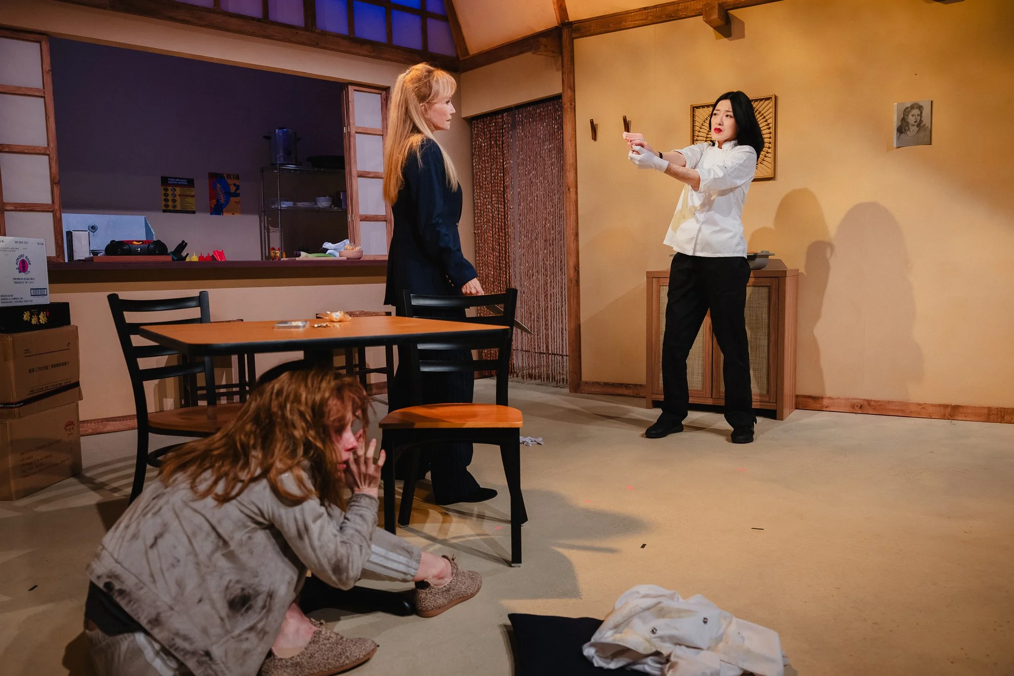 Three performers on a restaurant set: a blond woman in a navy blazer stands near the table, a red-haired woman crouches on the floor, and a dark-haired woman in a white chef's coat stands across the room pointing a gun