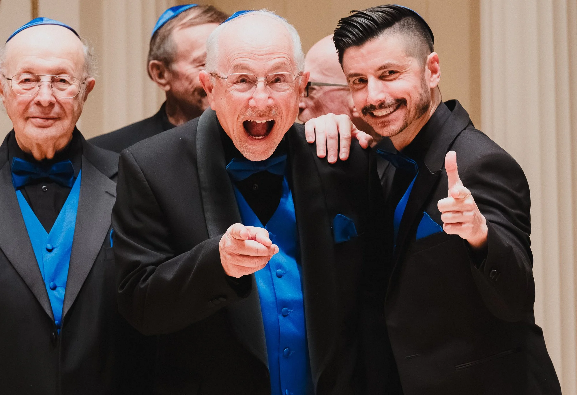 Two members of the San Diego Jewish Men's Choir sharing a laugh on stage at Weill Recital Hall, Carnegie Hall, one pointing at the camera and the other giving a thumbs up.