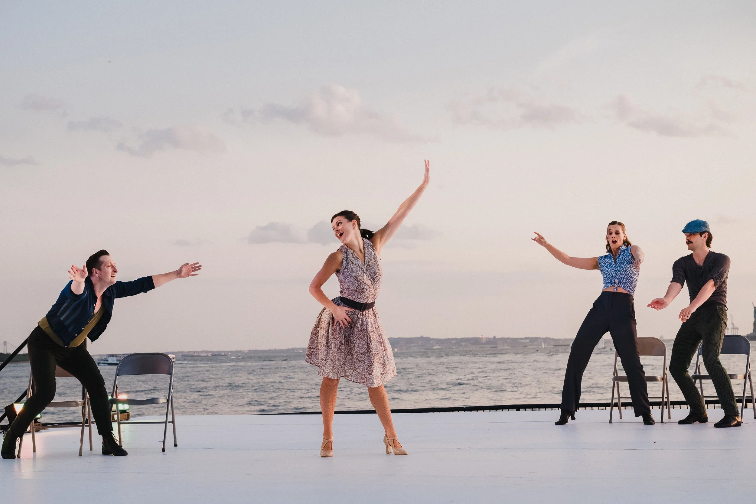 Four DPR Dance performers on the outdoor stage at Robert F. Wagner Jr. Park, arms raised wide, pale sky and harbor behind them