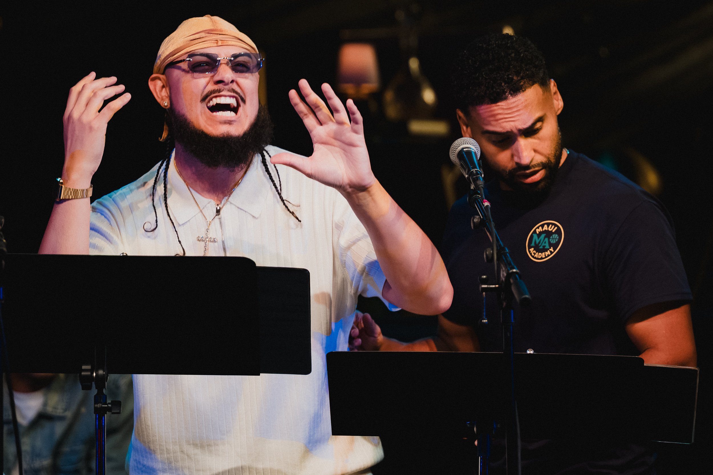 Fermin Suero, Jr. in a white shirt and orange head covering, arms raised and hands open mid-verse, at Greenwich House Theater