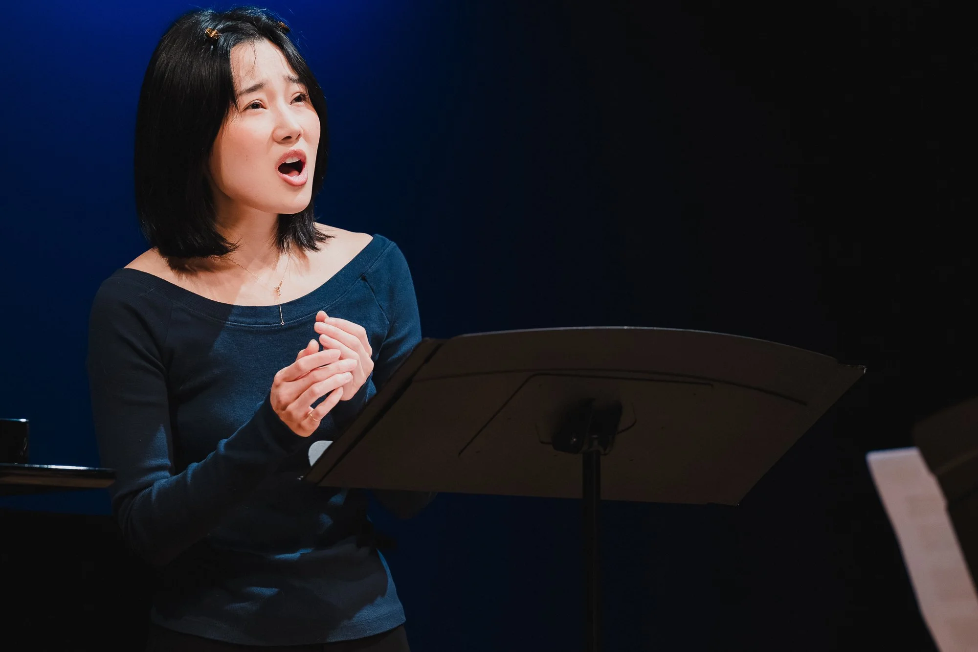 Soprano Katharine Li performing with hands clasped at her chest against a deep blue backdrop at Marc A. Scorca Hall