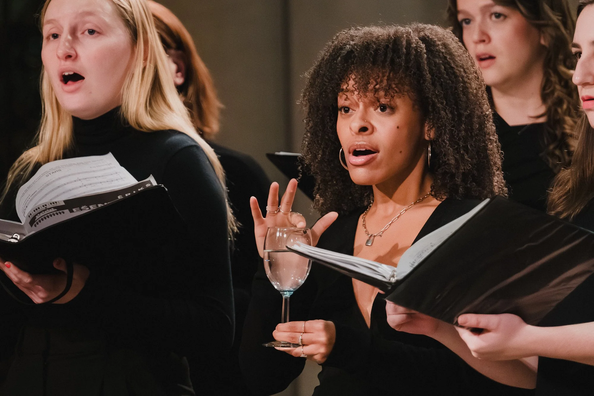 Treble singer with curly hair holding a tuned wine glass while singing, fellow singers with folders on either side.