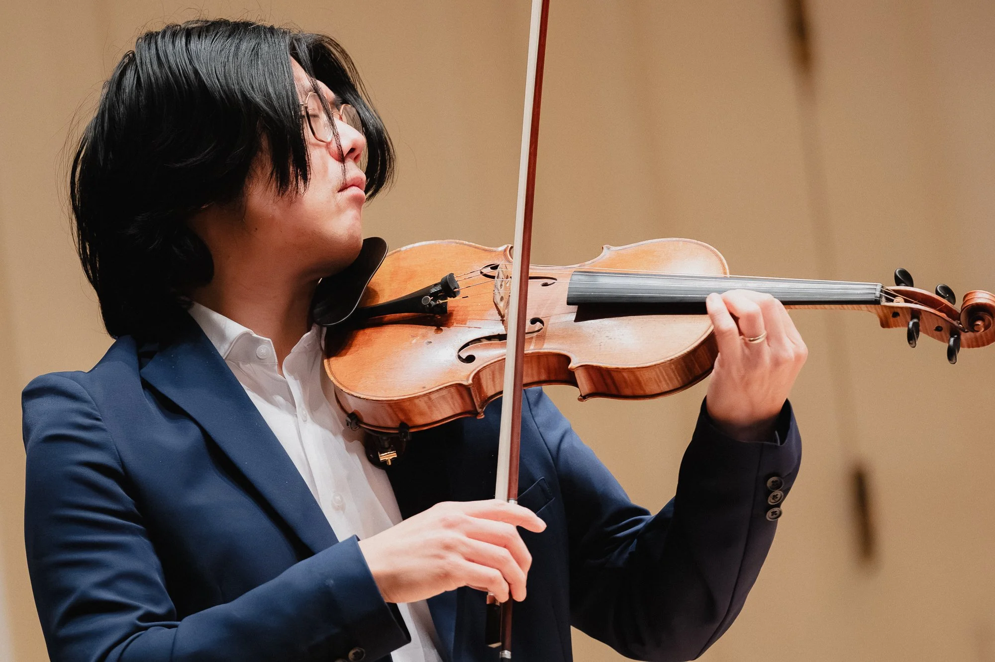 Young Asian male violinist in a navy suit and white shirt, eyes closed, holding the violin with bow arm extended, in front of a warm beige wall