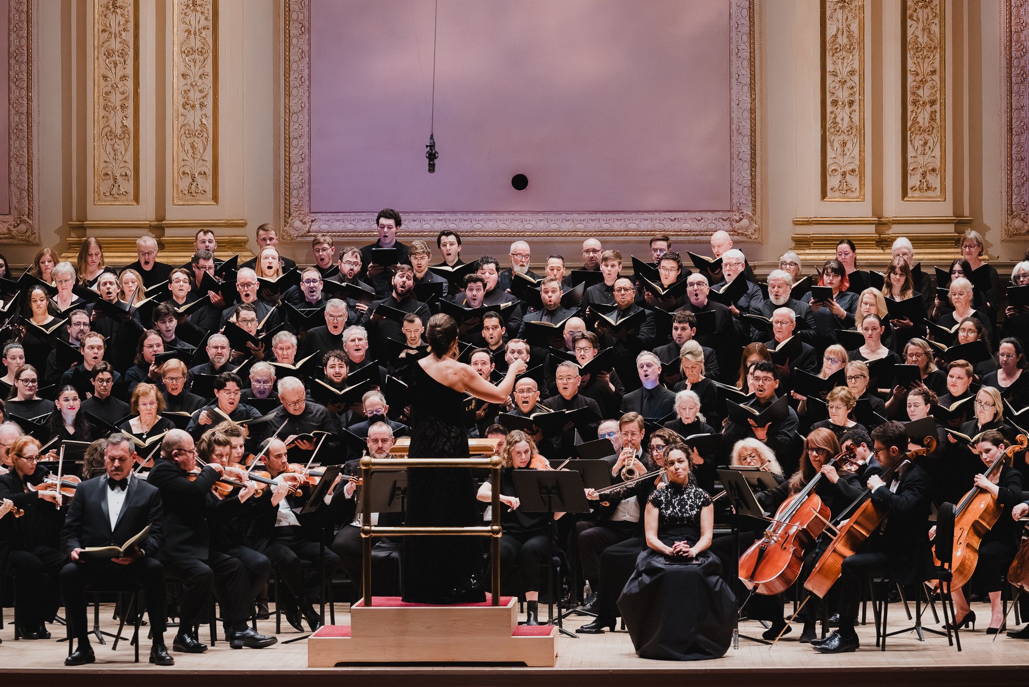 Female conductor seen from behind at the podium, full massed choir on risers with mouths open, pink-lit panel behind, orchestra in foreground