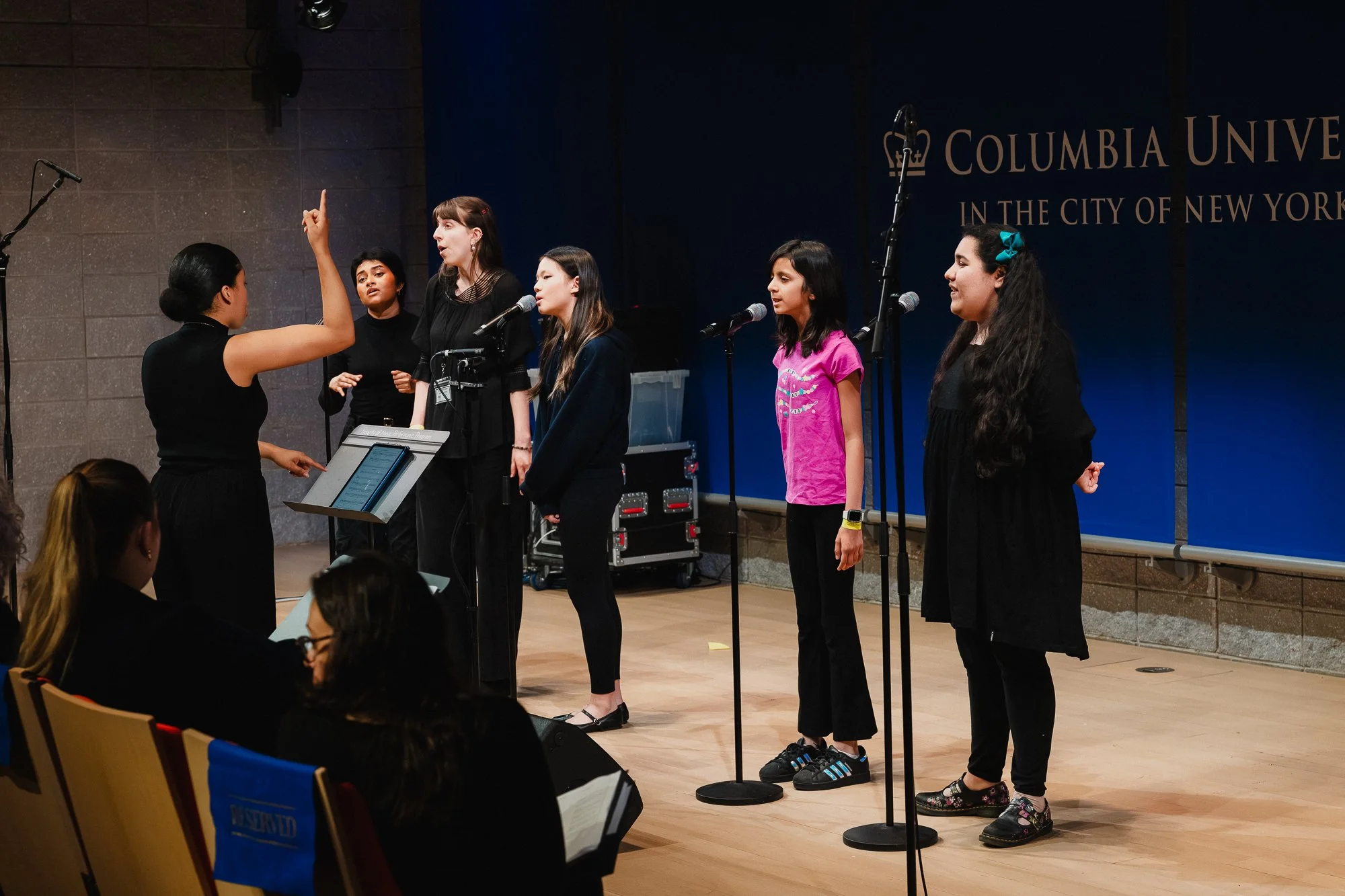 Small group of youth vocalists at microphone stands on stage, directed by a woman in black with one arm raised, Columbia University banner visible, one singer in pink shirt at right, viewed from house seats