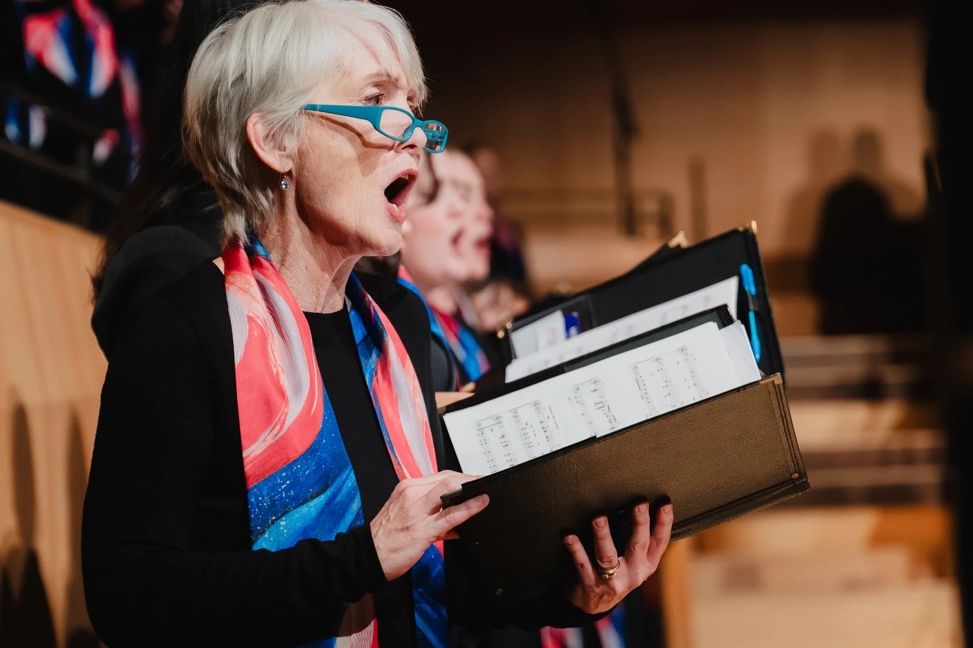Profile close-up of an older woman with gray hair and blue glasses, mouth open wide mid-phrase, holding a leather music binder with sheet music, another singer visible slightly behind her