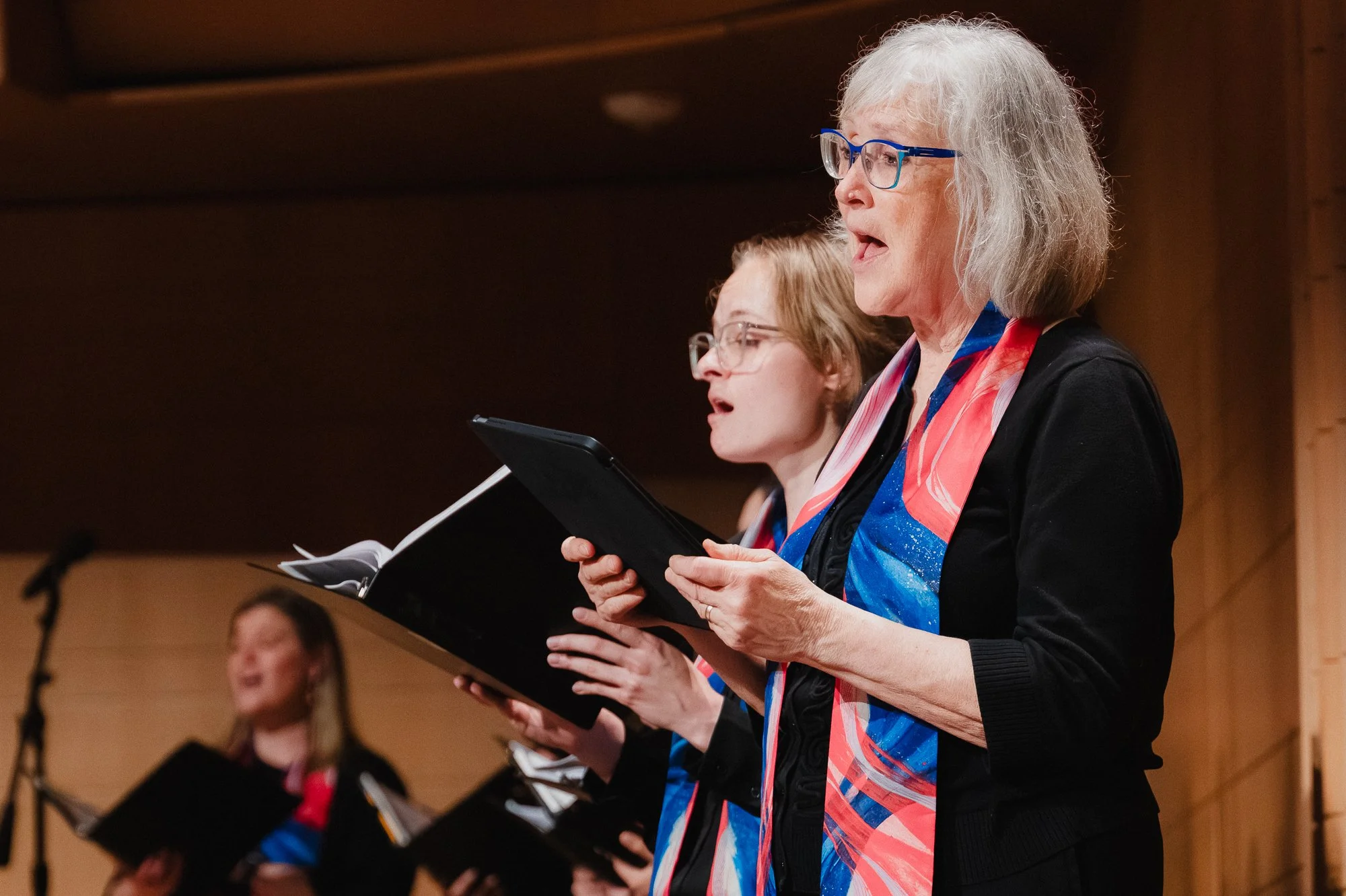 Two singers in profile, both mouths open singing, wearing black with blue, pink, and red scarves, holding black folders, warm wooden acoustic panels in the background, a younger blonde singer and an older gray-haired singer side by side