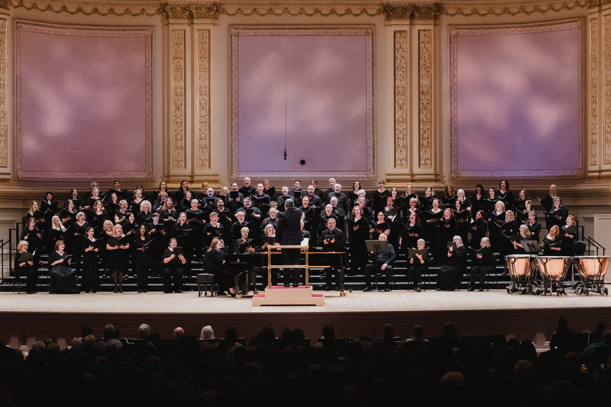 Large massed choir singing on risers behind a conductor seen from below and behind, three pink-lit panels on the Stern Auditorium back wall, timpani visible at right
