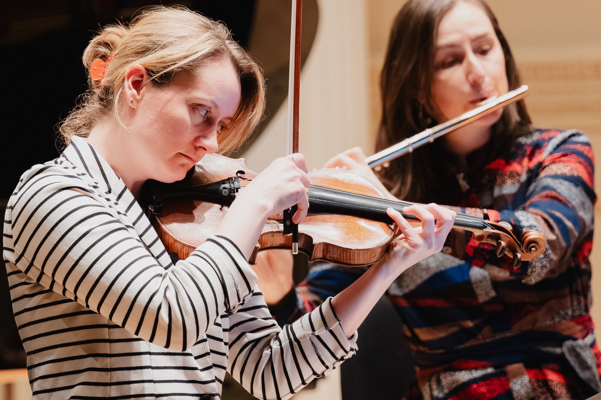 Two violinists in close-up, one in soft focus in the foreground in a patterned top, the other in sharp focus behind her in a striped shirt, on the stage of Weill Recital Hall