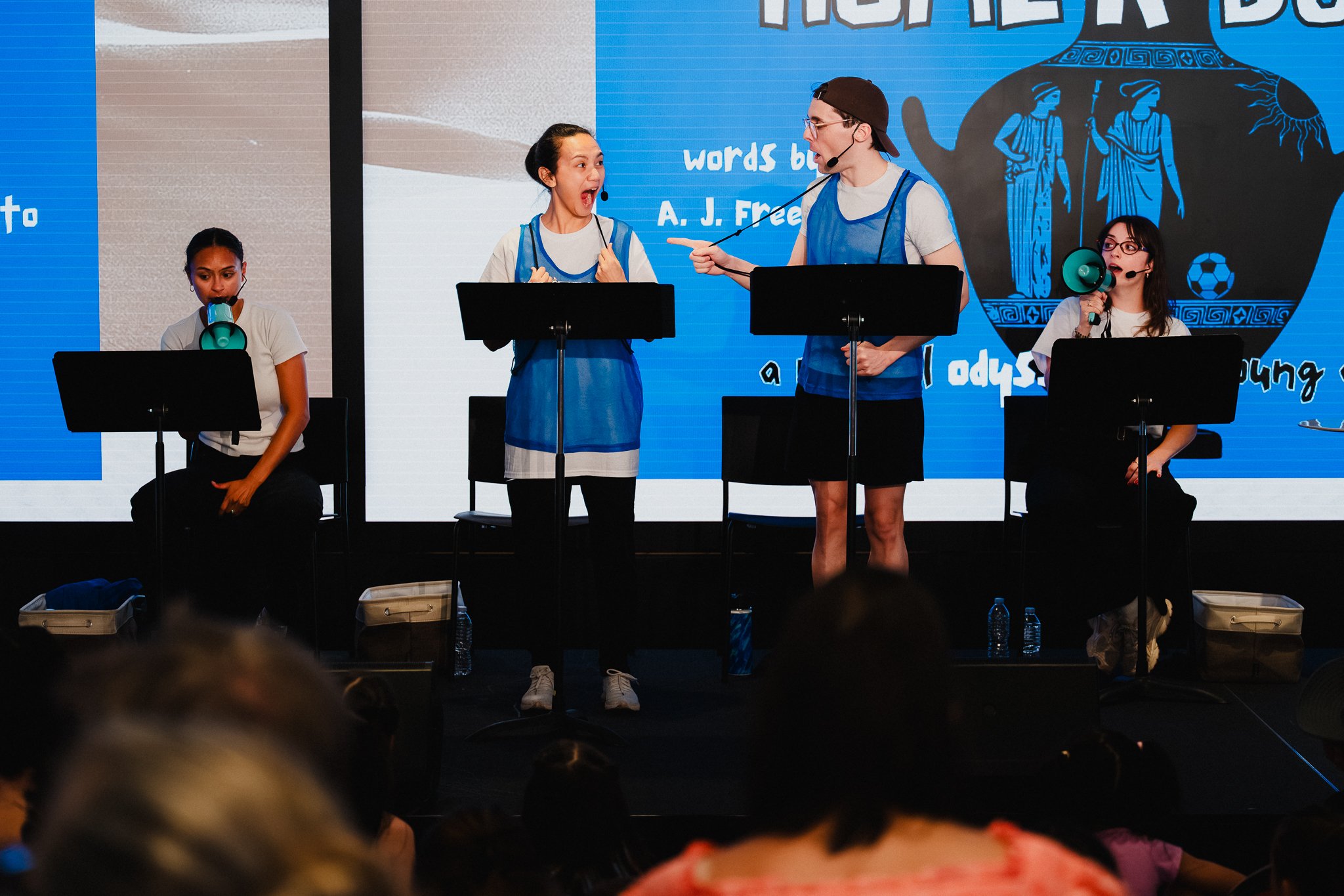 Four performers at music stands on a low stage, one holding a green megaphone, a title card reading 'Home'r Bust!' on a screen behind them, audience heads in the foreground