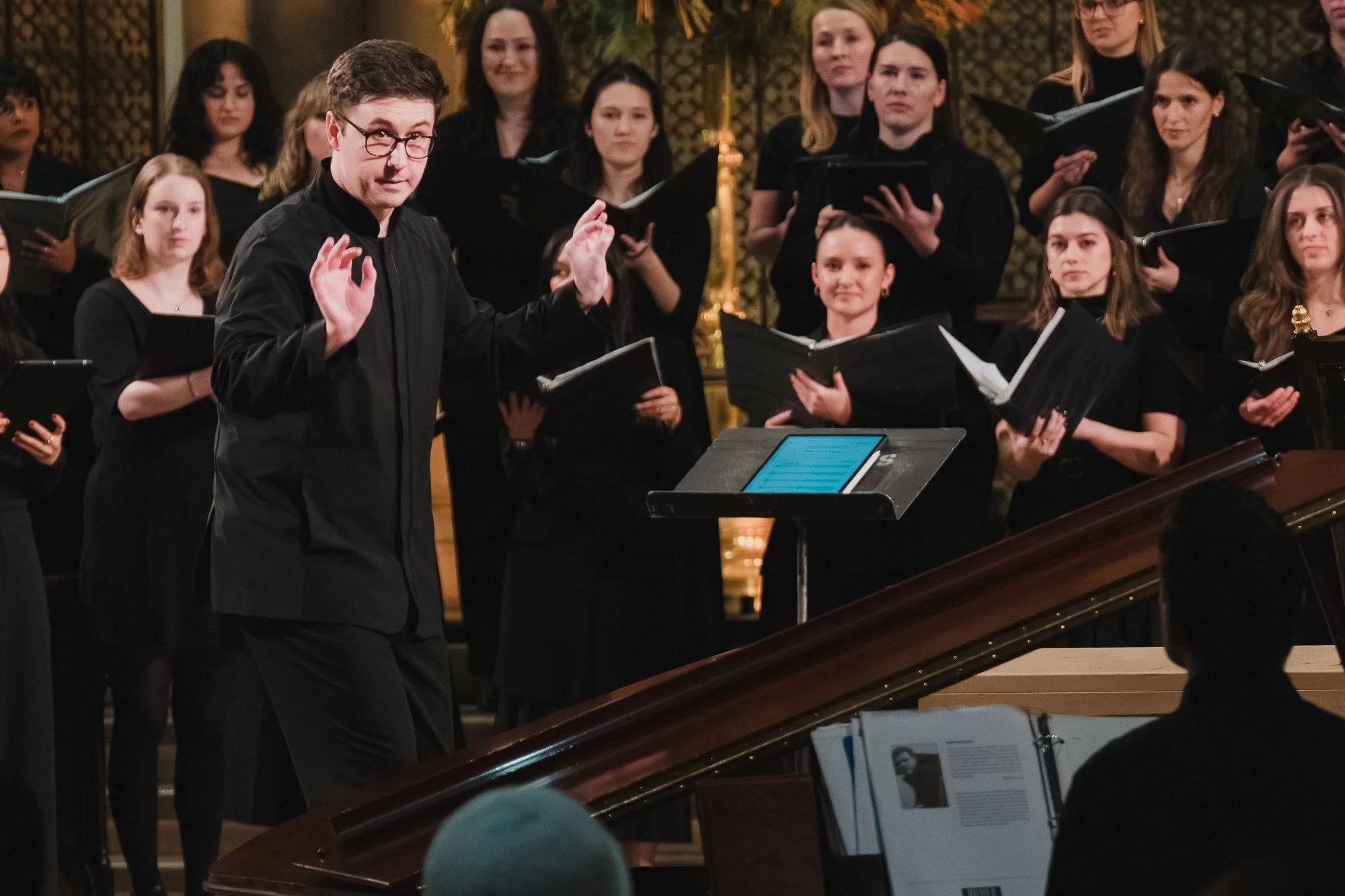 Artistic director Alex Canovas conducting with expressive hands, choir and grand piano visible, at the Church of St. Mary the Virgin.