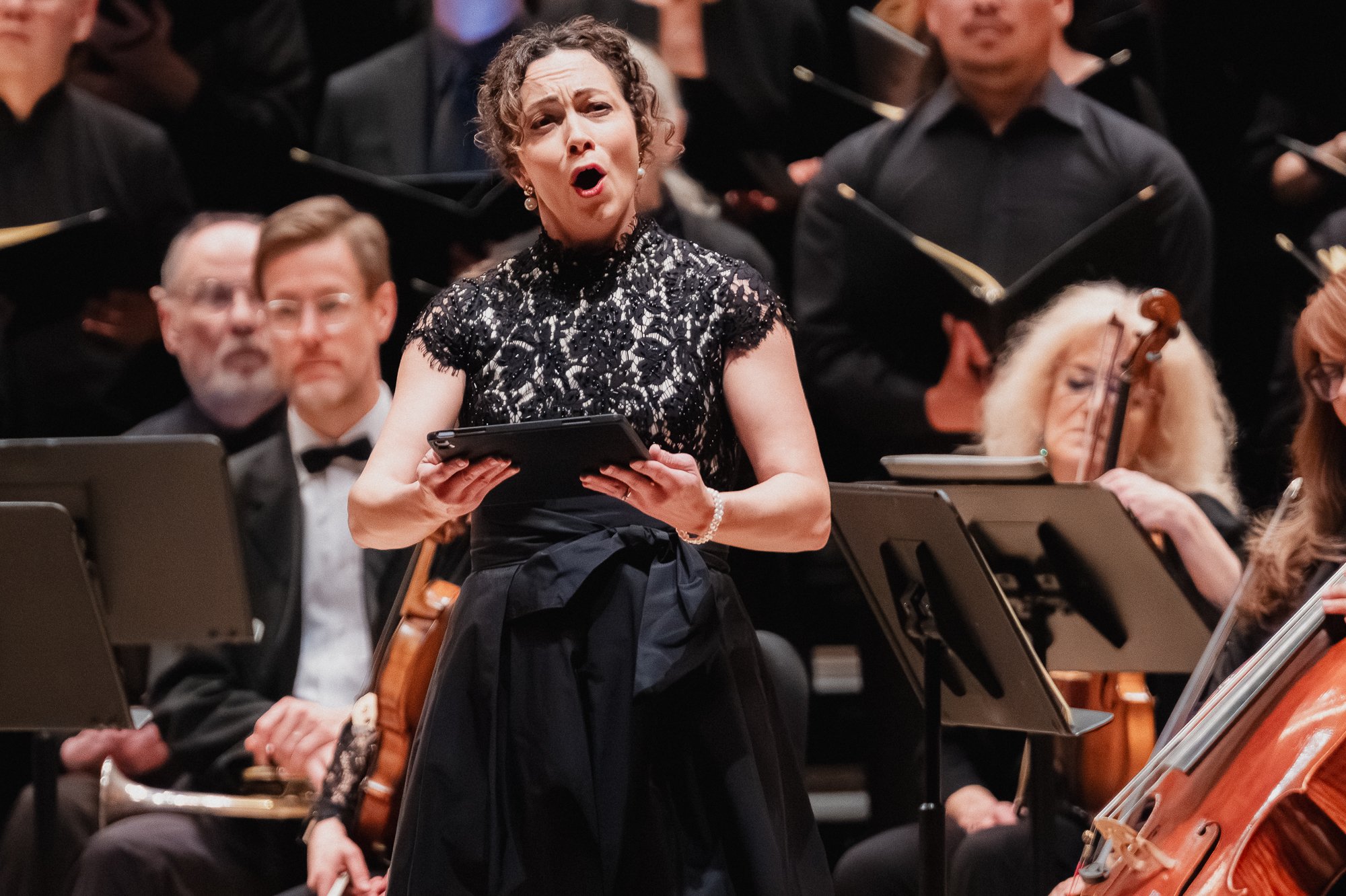 Mezzo-soprano in black lace top and full skirt singing with mouth open and chin raised, holding a tablet, orchestra and chorus visible behind her