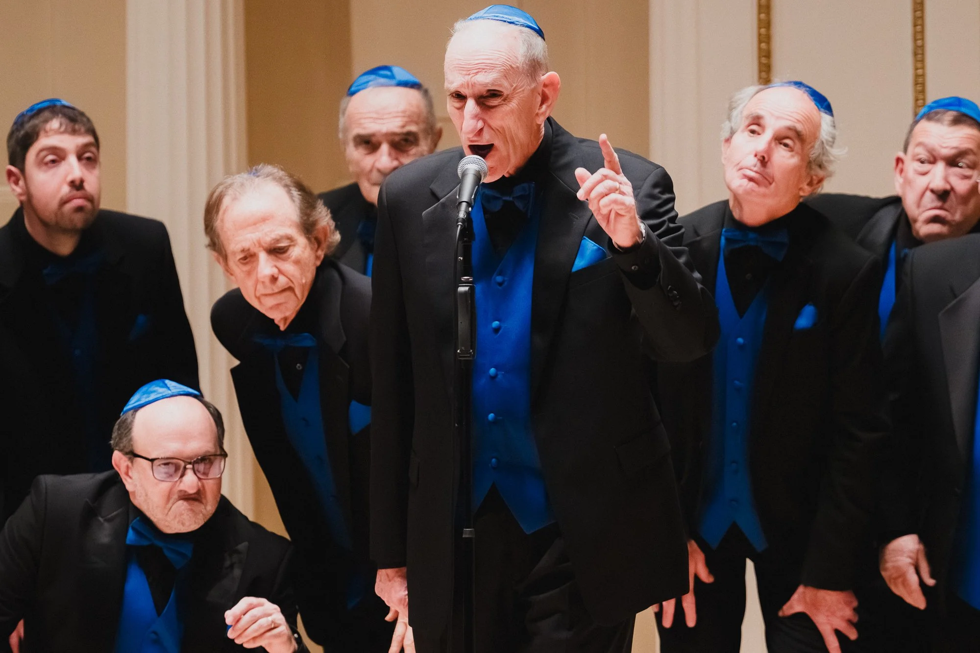 A soloist performing at the microphone with the San Diego Jewish Men's Choir at Weill Recital Hall, Carnegie Hall, surrounded by expressive faces from fellow choir members.