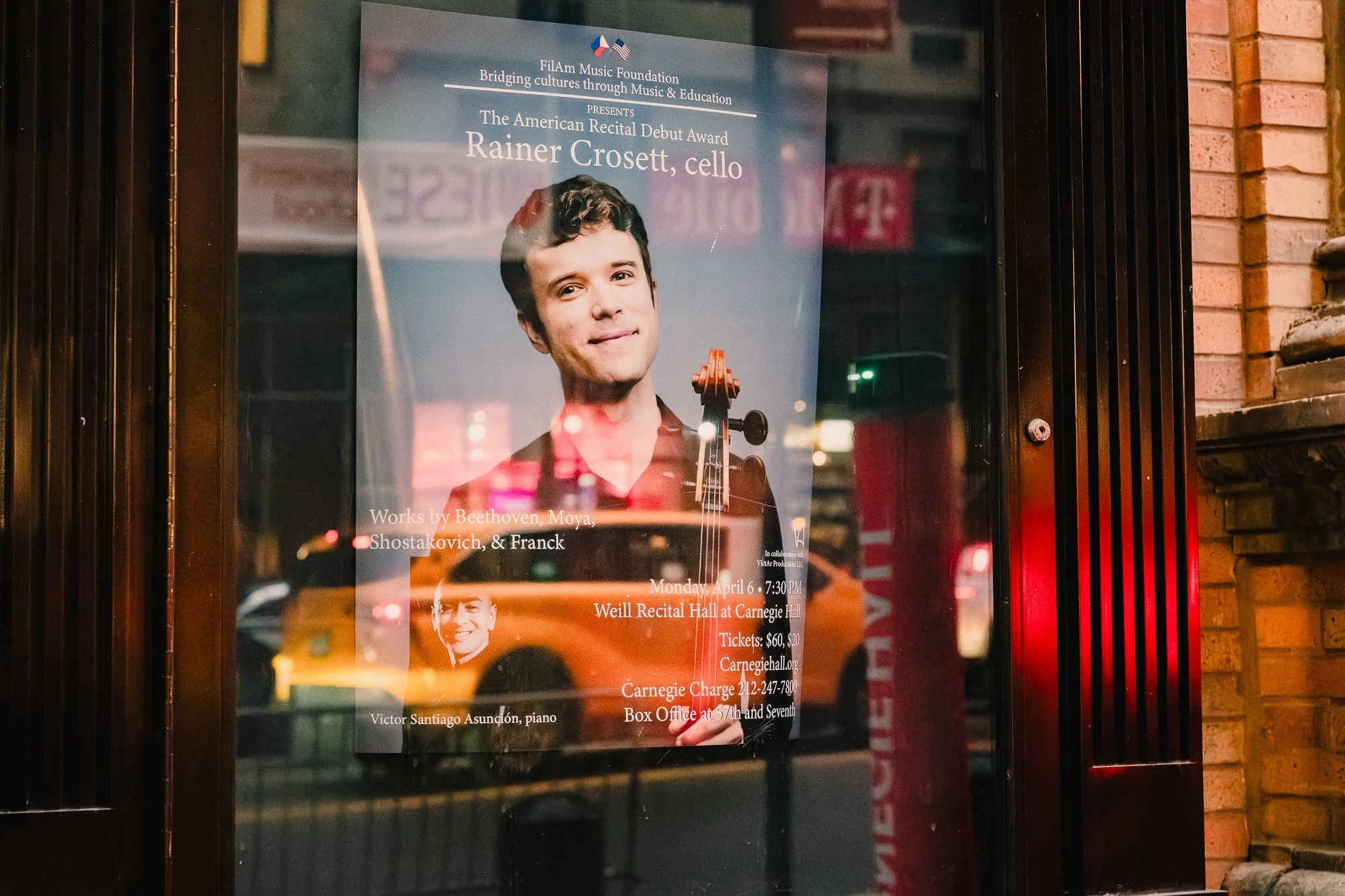 Recital poster for cellist Rainer Crosett's American Recital Debut Award   concert displayed in a Carnegie Hall vitrine at dusk, glass reflecting a         passing yellow taxi and city lights against the brick facade.