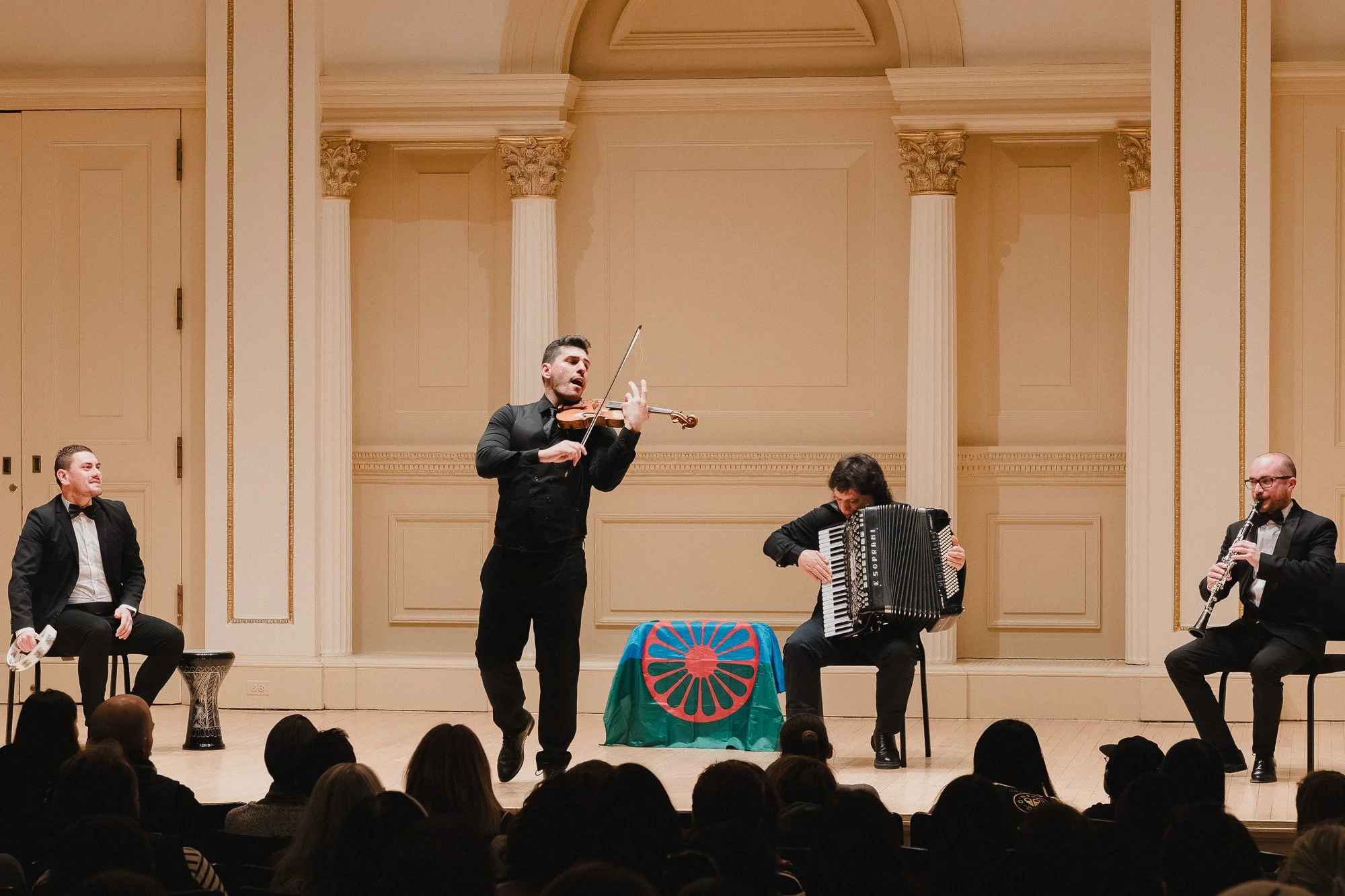 Four musicians performing on stage at Weill Recital Hall: percussionist, violinist, fisarmonica player, and clarinetist, with Romani flag displayed center stage
