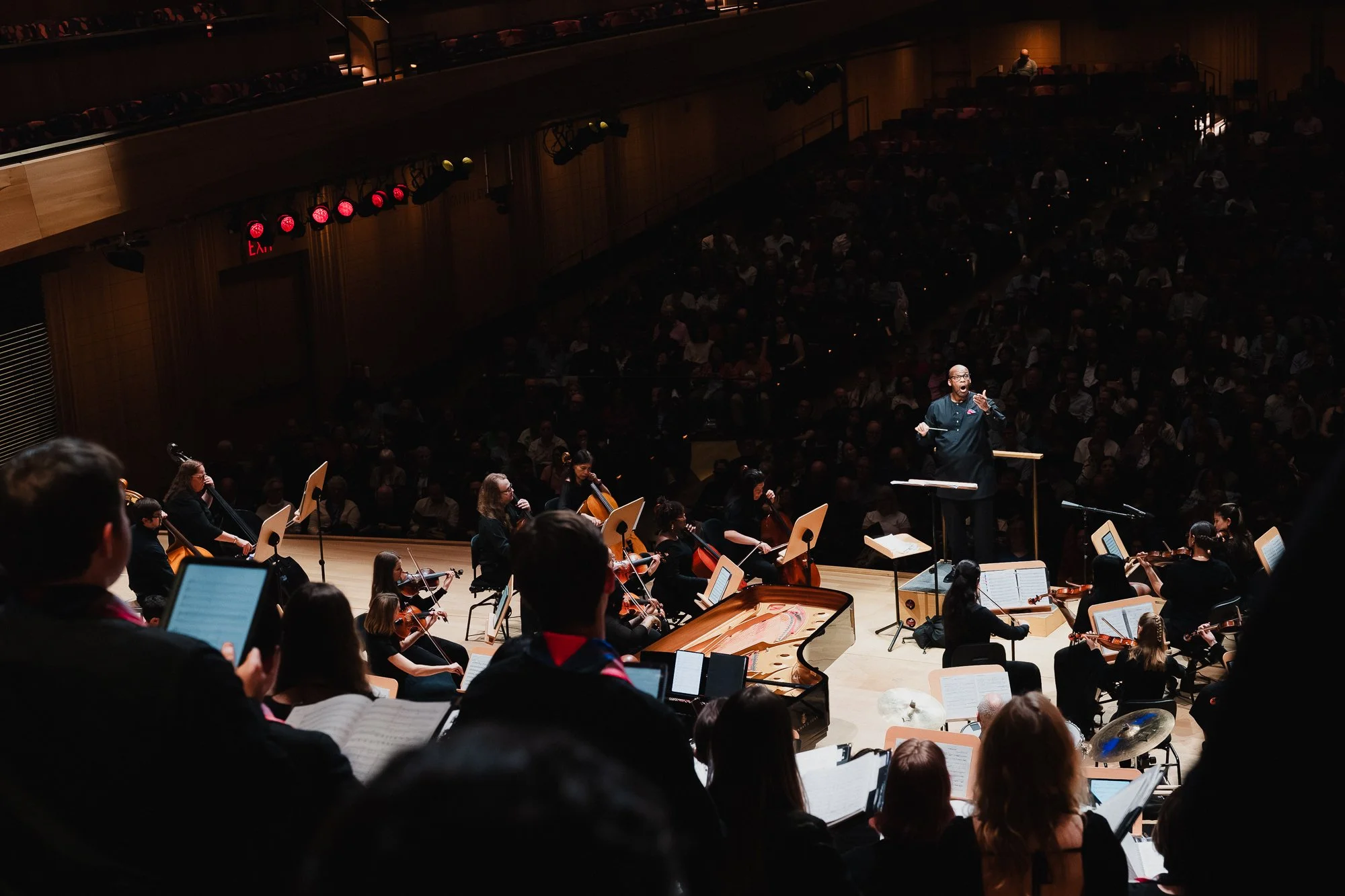 Overhead view from upper balcony looking down at the orchestra on stage, conductor at podium with arms raised addressing the audience, a grand piano visible center stage, choir members visible at the lower edge of the frame
