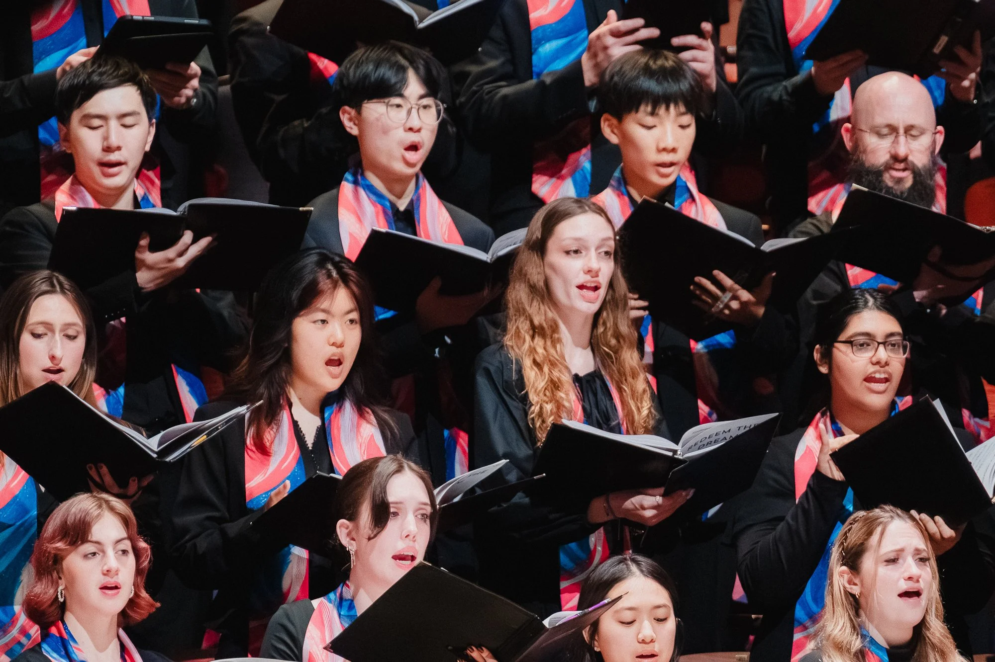 Close shot of a row of choir singers in black with colorful scarves, several mouths open mid-phrase, holding black music folders, a mix of young singers visible, one folder showing text reading "Redeem the Dream"