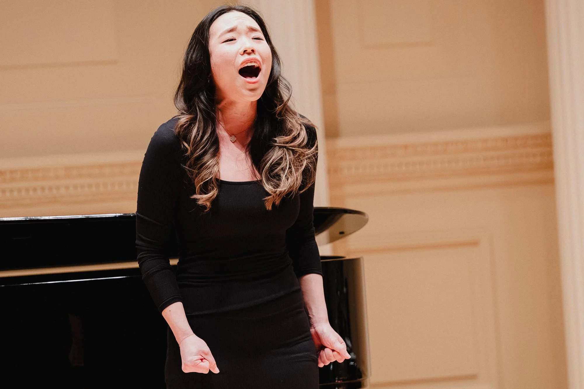 A woman in a black long-sleeve dress sings with both fists clenched at her sides, head tilted back, mouth fully open, beside a black grand piano
