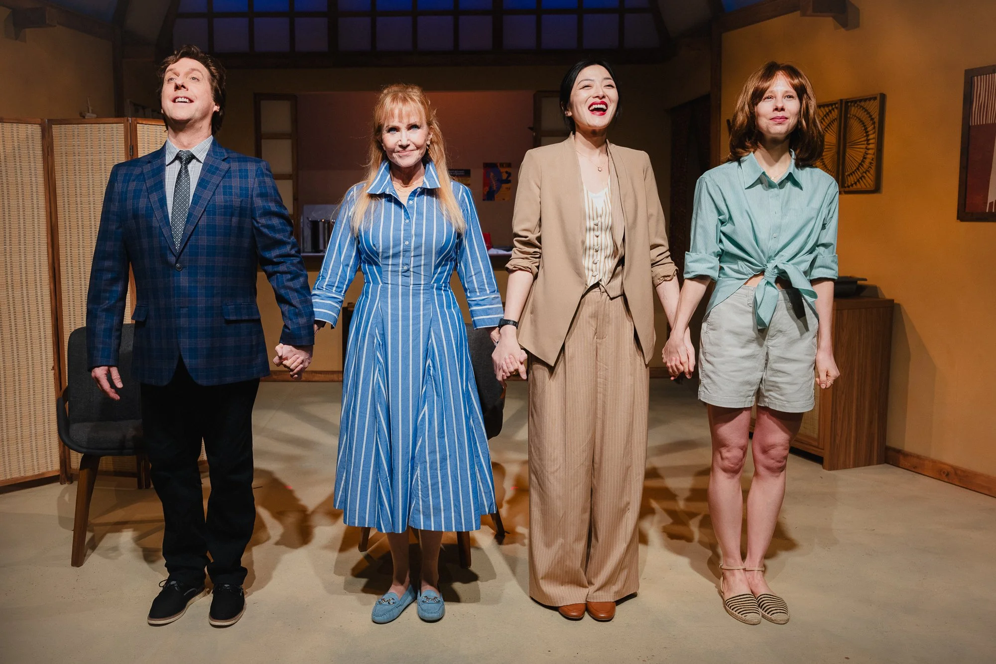 Four actors stand in a line holding hands for a curtain call bow on a warmly lit stage: a man in a blue plaid blazer, a woman in a blue striped dress, a woman in a tan blazer laughing, a woman in a teal shirt and shorts