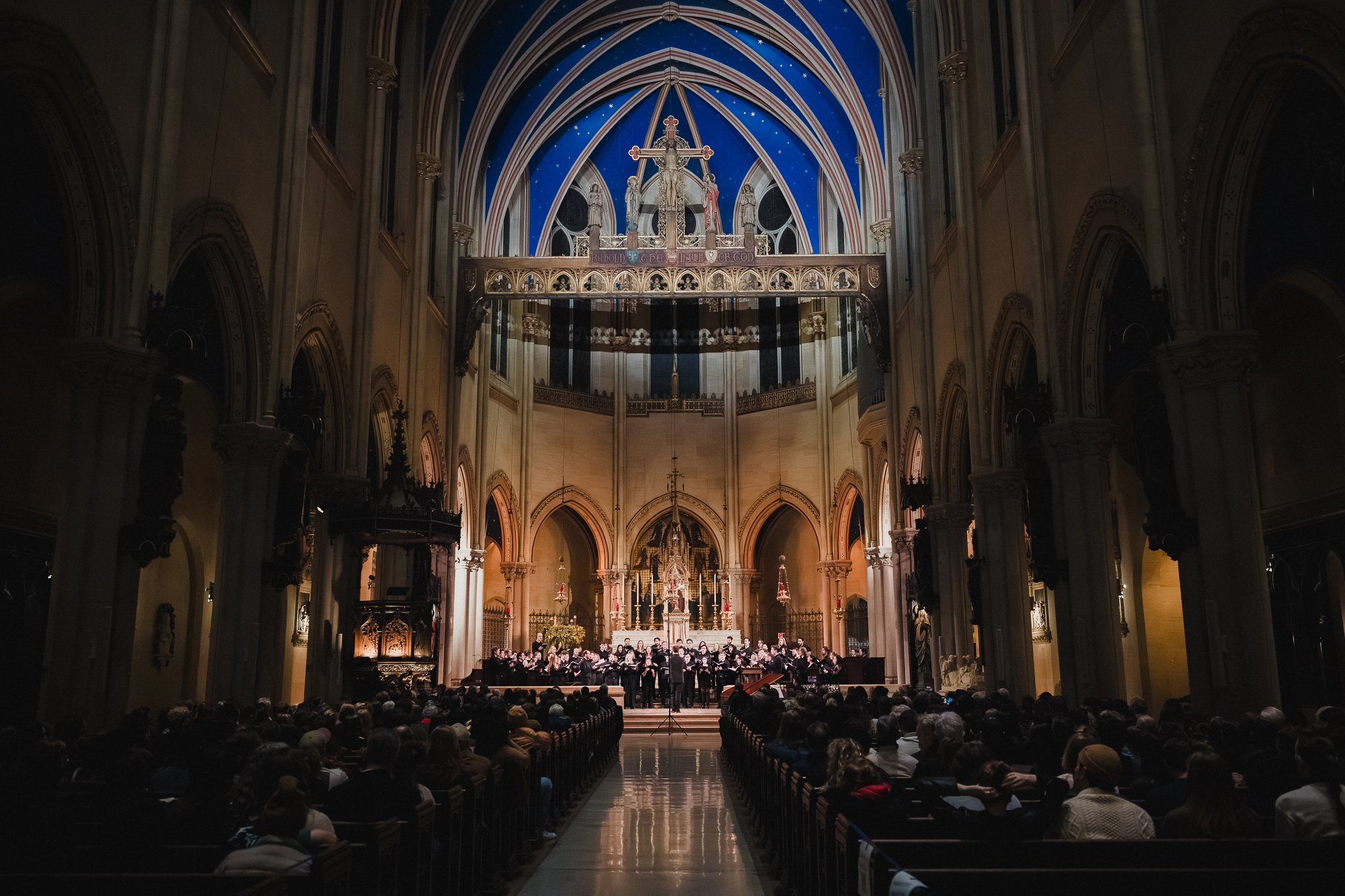 Wide shot of the Church of St. Mary the Virgin sanctuary showing full audience in pews, choir performing at altar, blue vaulted ceiling with stars visible above.