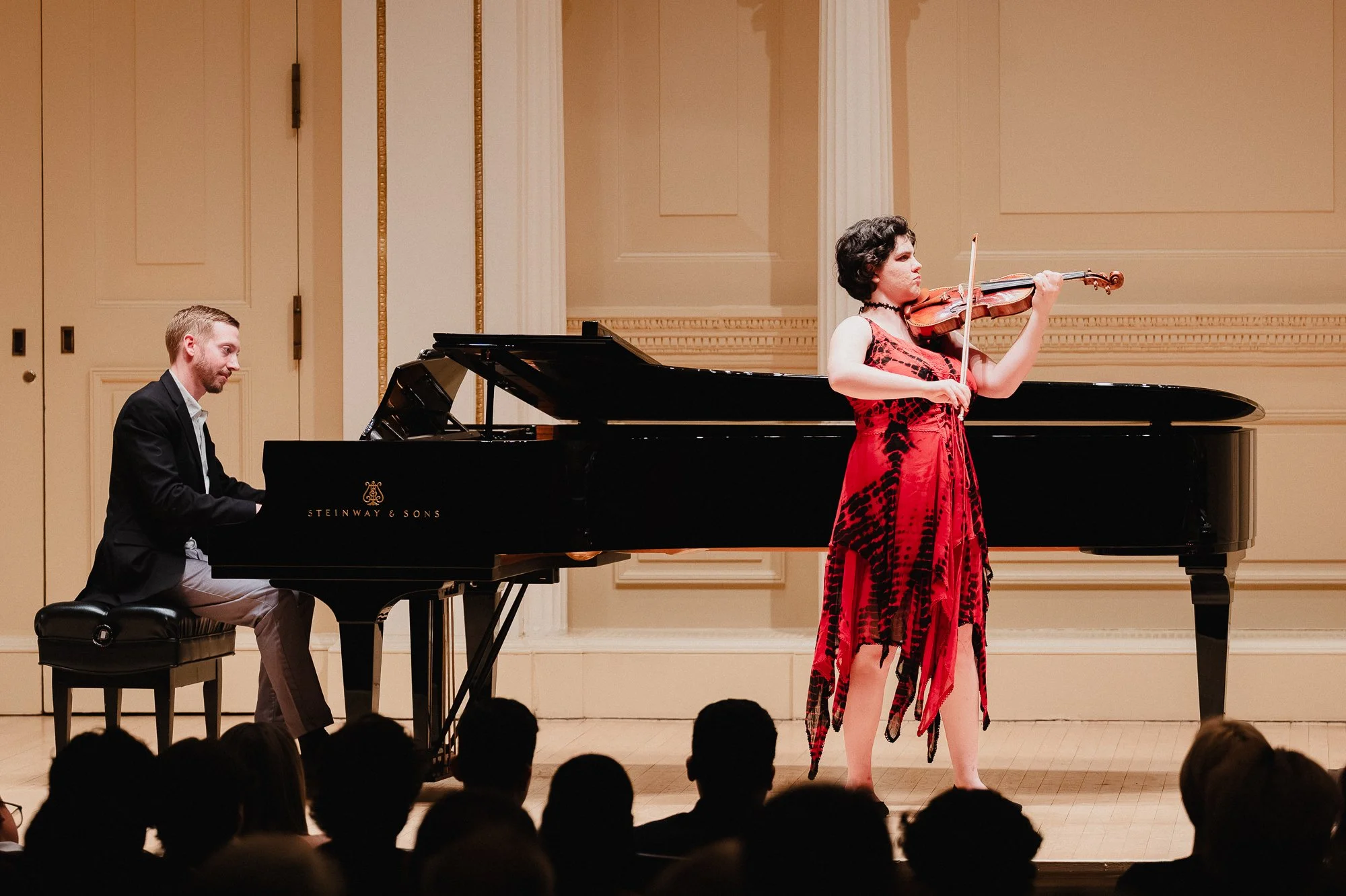 A girl in a flowing red-and-black dress plays violin center stage, bow extended, barefoot, with a man at a Steinway piano visible to the left and audience silhouettes in the foreground