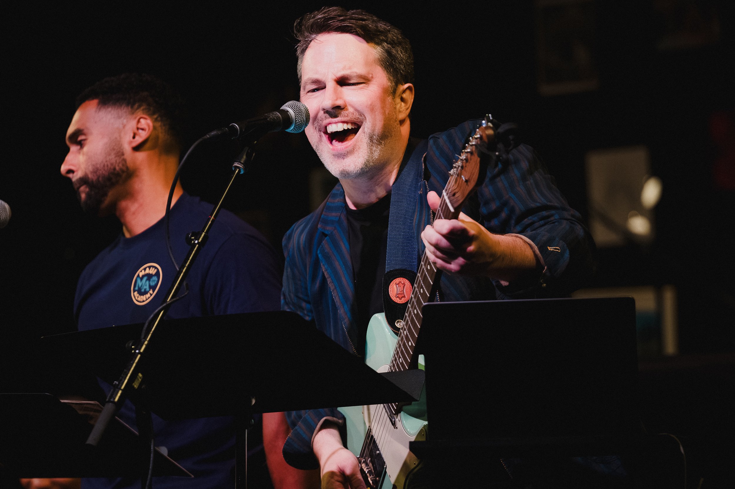 Pete White sings into a mic while holding his pale green guitar at Greenwich House Theater, another performer partly visible at left