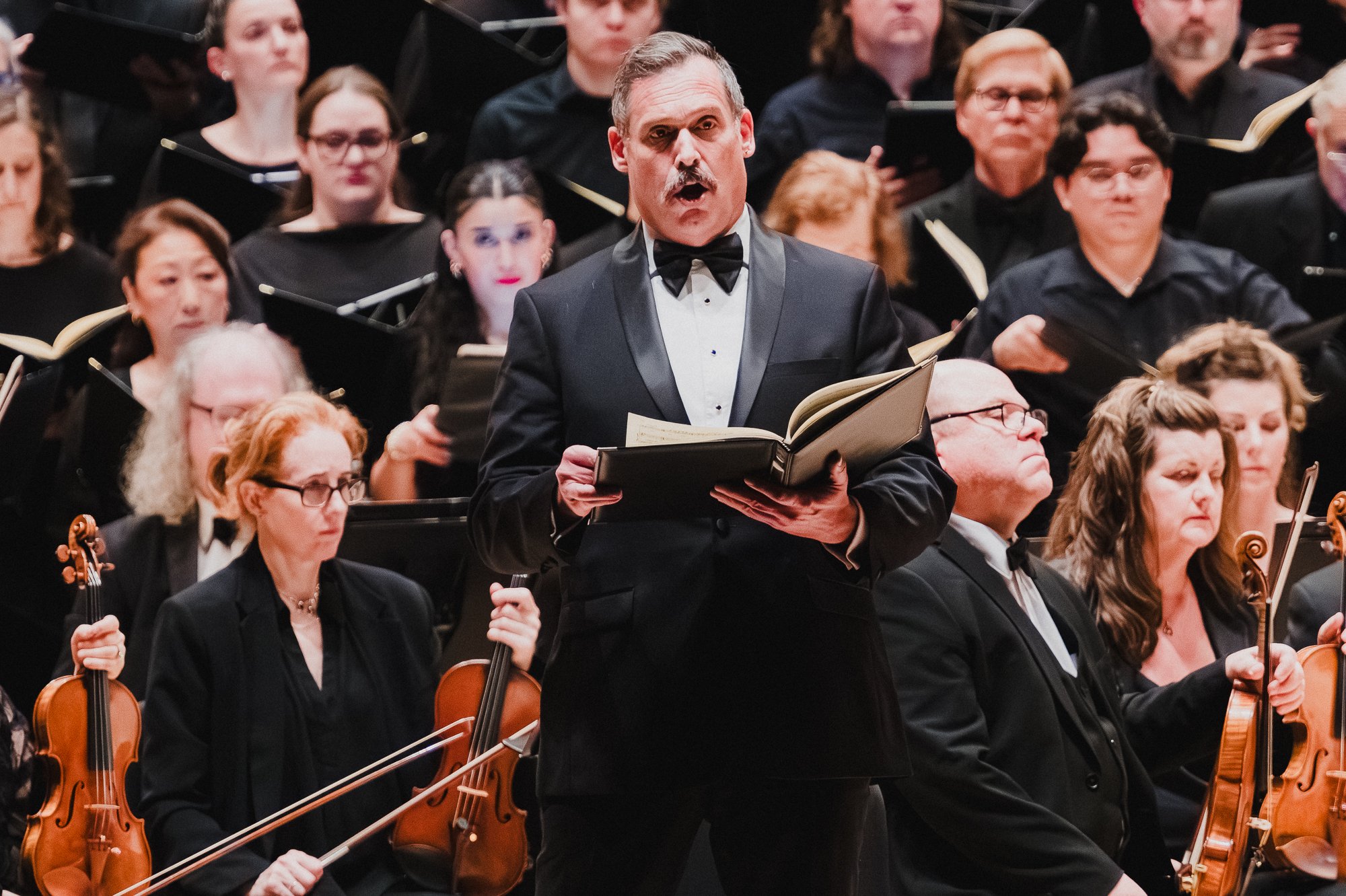 Baritone in black tie holding an open score, singing, surrounded by seated violin players and choral singers on risers behind